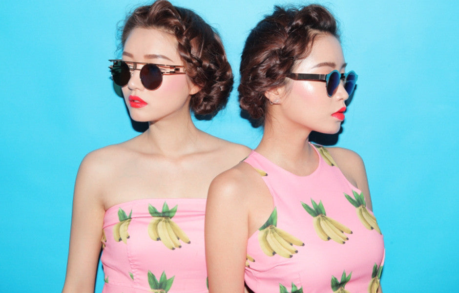 Stylish photo of two models in sunglasses and bright makeup against a blue background, one wearing a banana-print top.