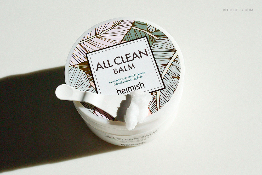 A top-down shot of a white tub of Heimish All Clean Balm. The lid features a tropical leaf pattern, and a small amount of the white balm product is visible on the spatula resting on the tub.