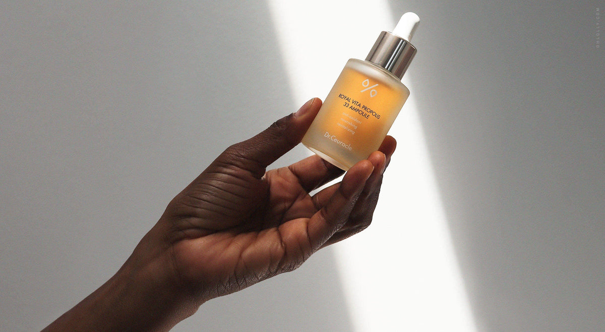 A person with dark skin holds a clear glass bottle of Dr. Ceuracle Royal Vita Propolis 33 Ampoule against a backdrop of bright, direct sunlight.