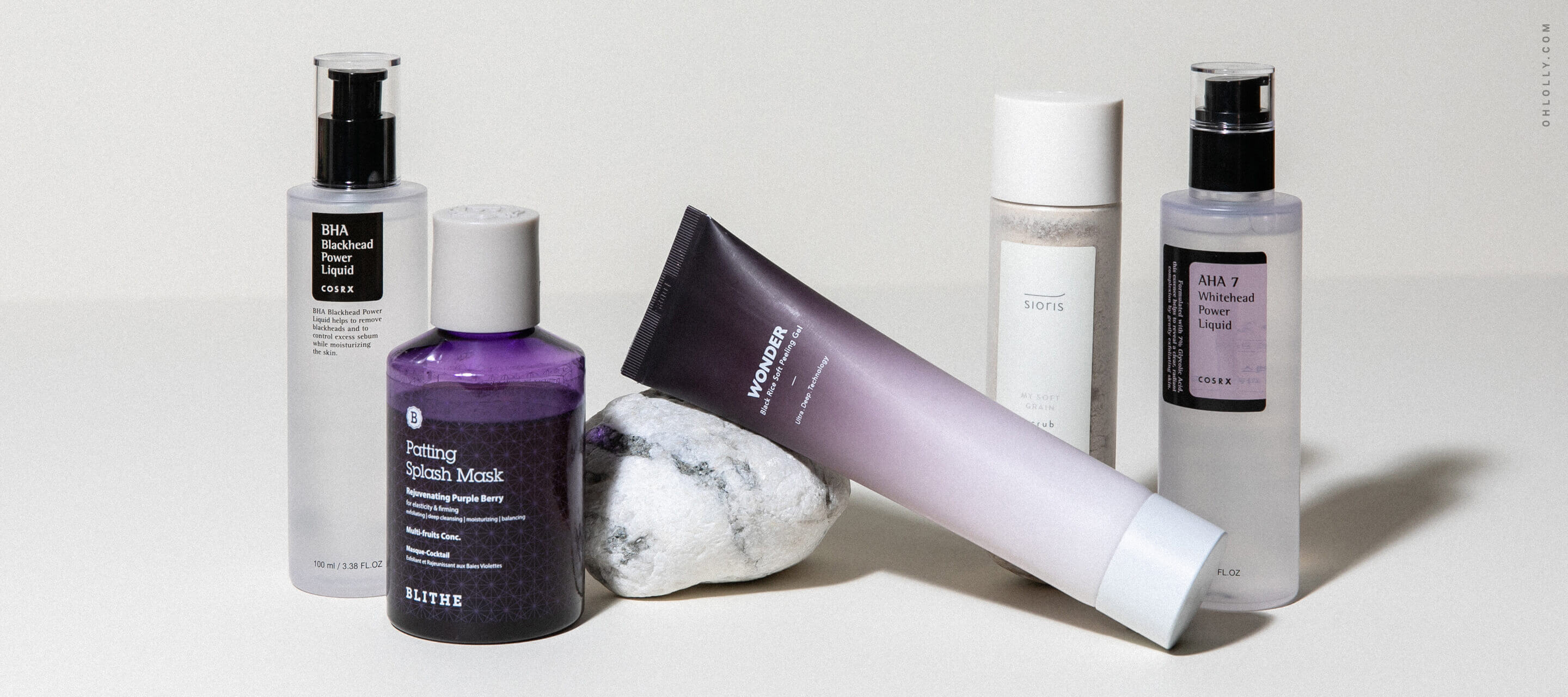 Various skincare products, including a purple jar, a gradient tube, and several tall clear bottles, are arranged around a white rock on a plain background.