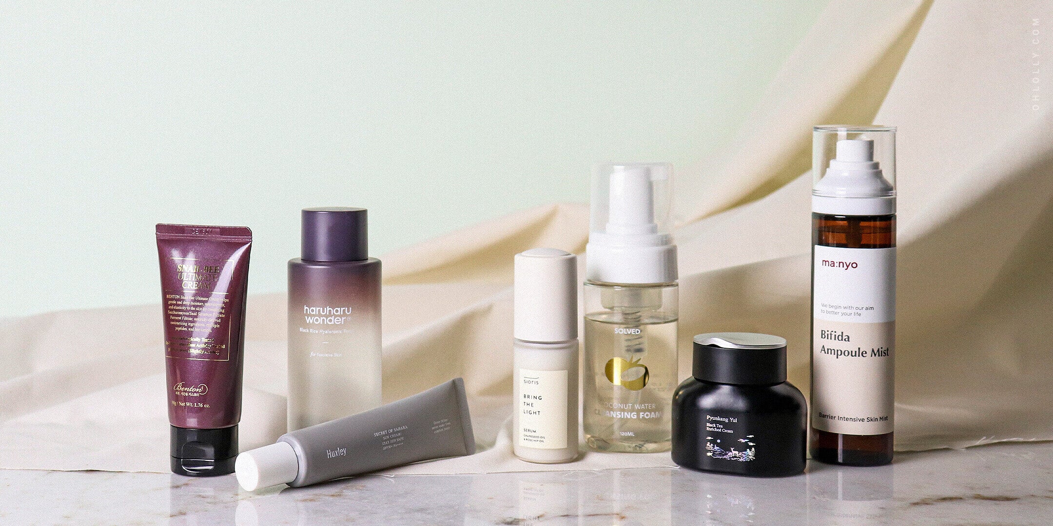 An assortment of various K-beauty skincare bottles and tubes, including creams and serums, arranged on a marble surface with draped fabric in the background.