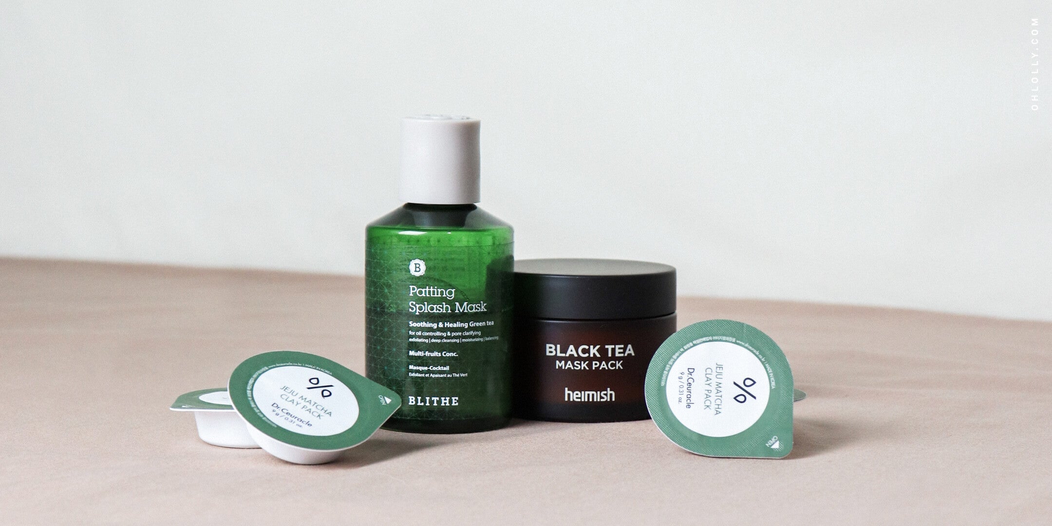 Four skincare products, including a green glass bottle of BLITHE Patting Splash Mask and a black jar of Heimish Black Tea Mask Pack, on a neutral surface.