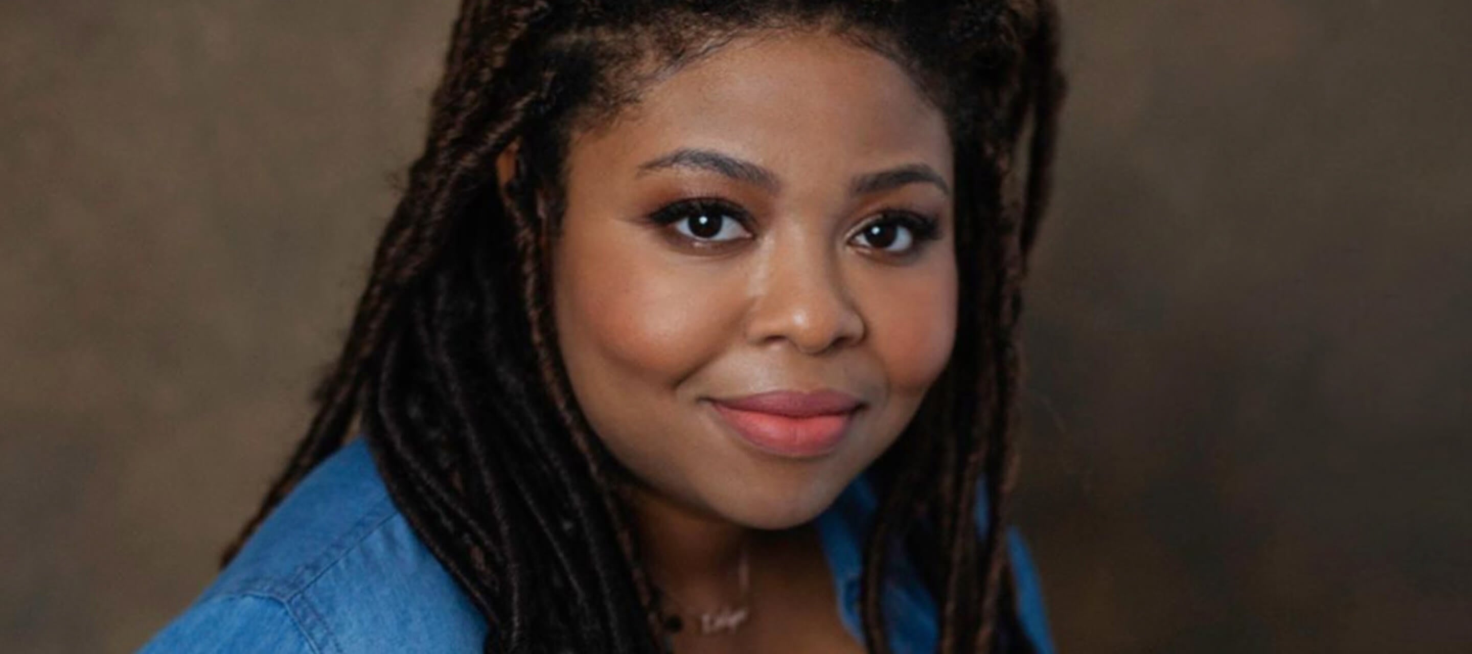 A professional headshot of a woman with dark, braided hair looking at the camera with a slight smile while wearing a blue denim shirt.