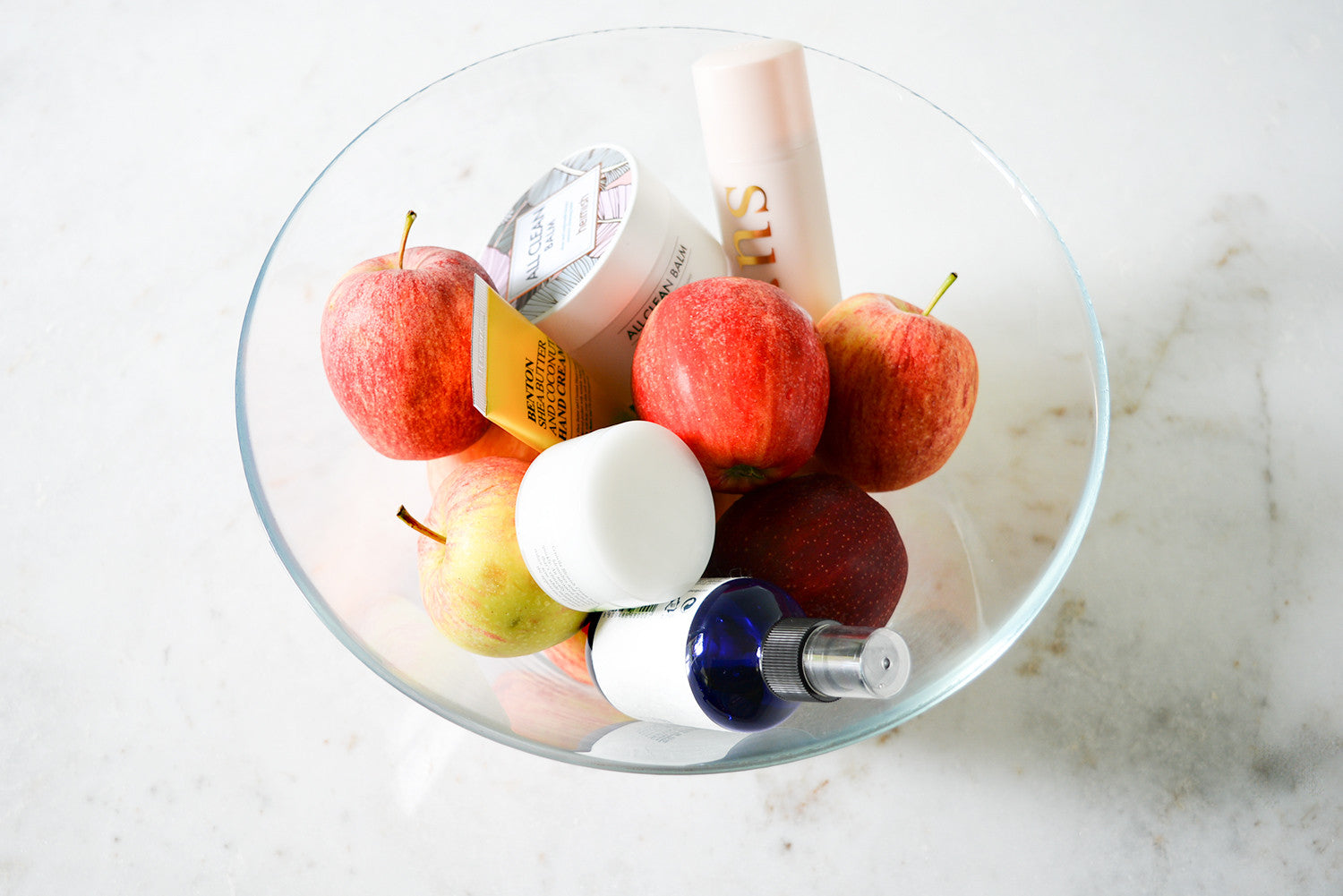 A clear glass bowl containing several red apples and various small skincare products, including a white jar and a blue dropper bottle.