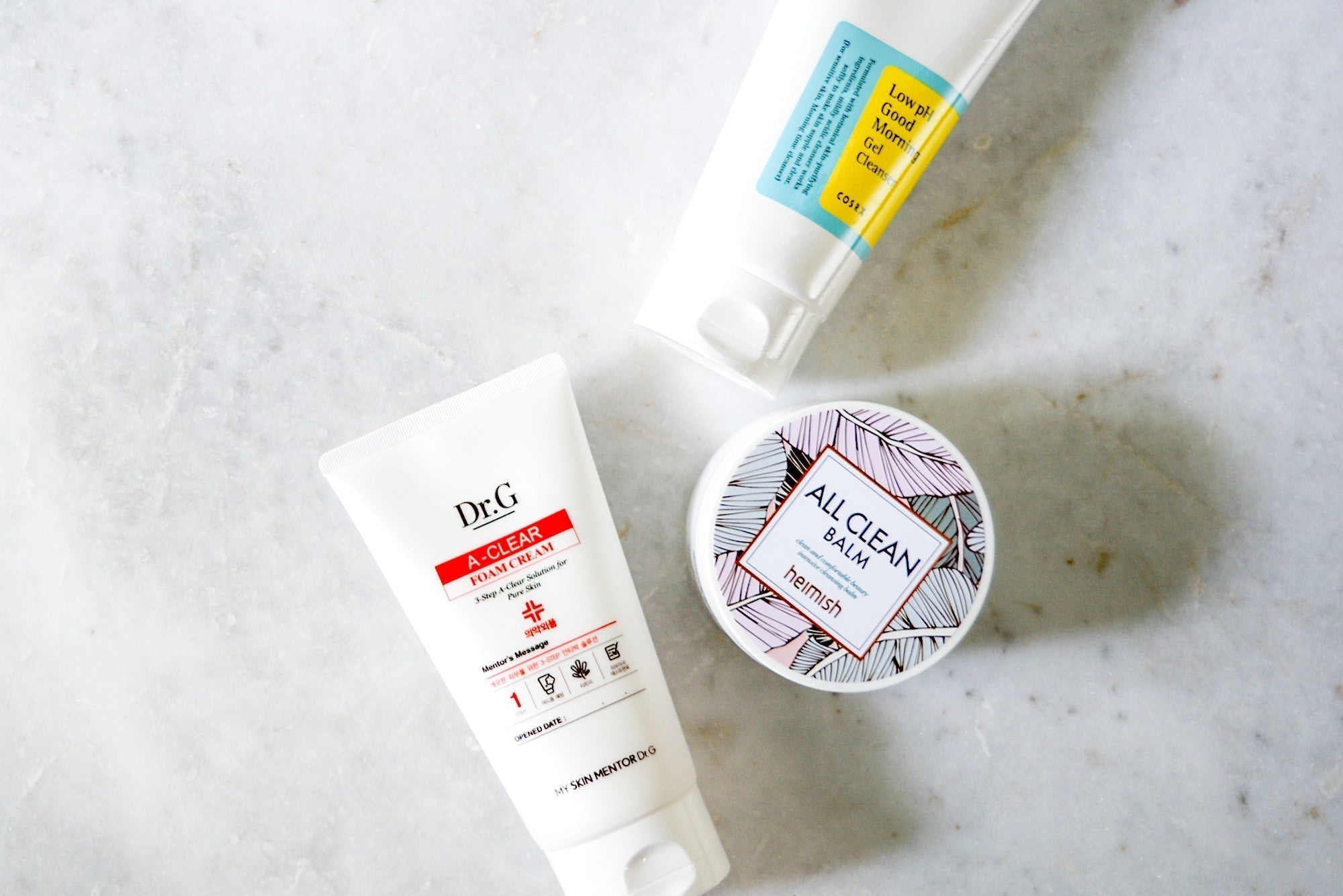 Flat lay photo of three skincare products on a white surface: a white tube with red and black text, a blue and yellow tube, and a round, patterned container.