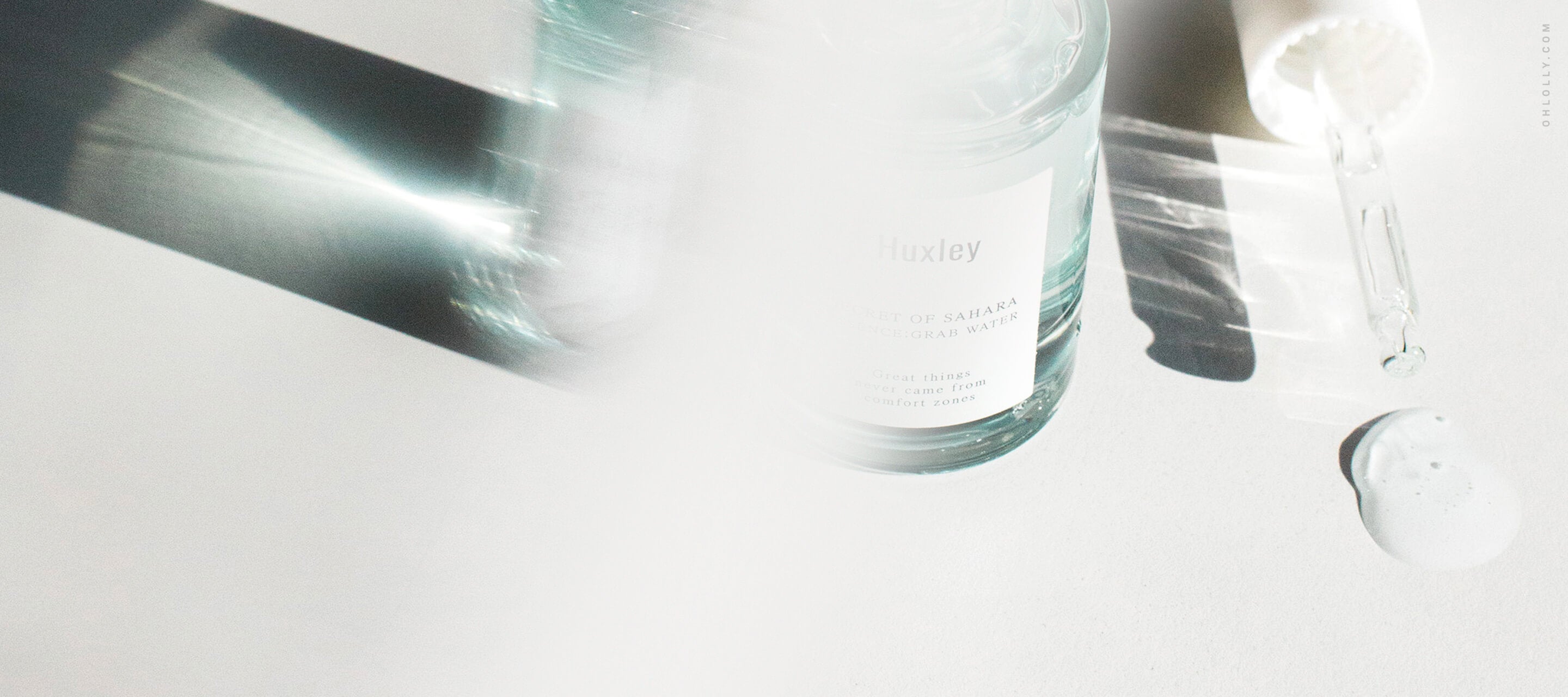 A glass skincare bottle with a dropper and a small amount of clear liquid on a white surface, shown with a bright, blurry light flare.