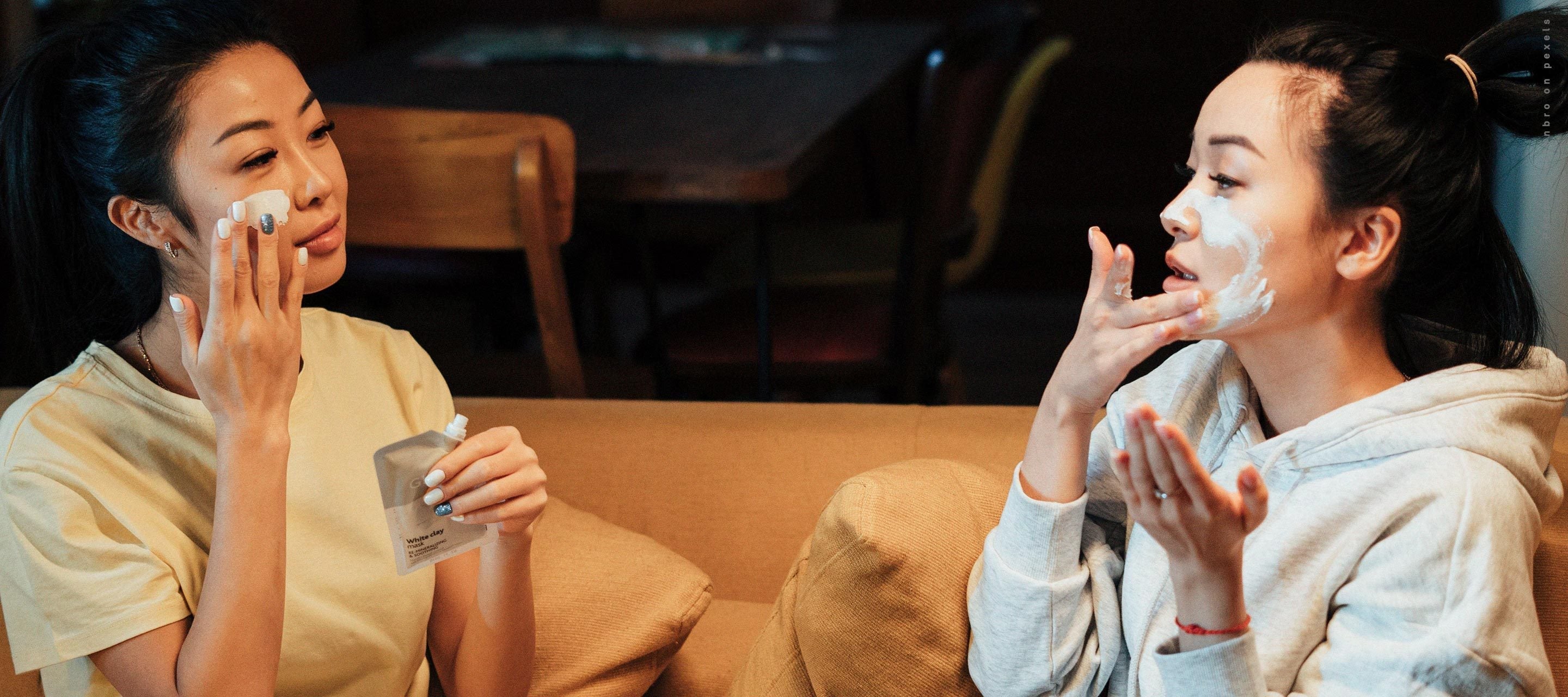 Two young women sitting on a couch, applying white face masks to their cheeks and foreheads as they enjoy a relaxing night in.