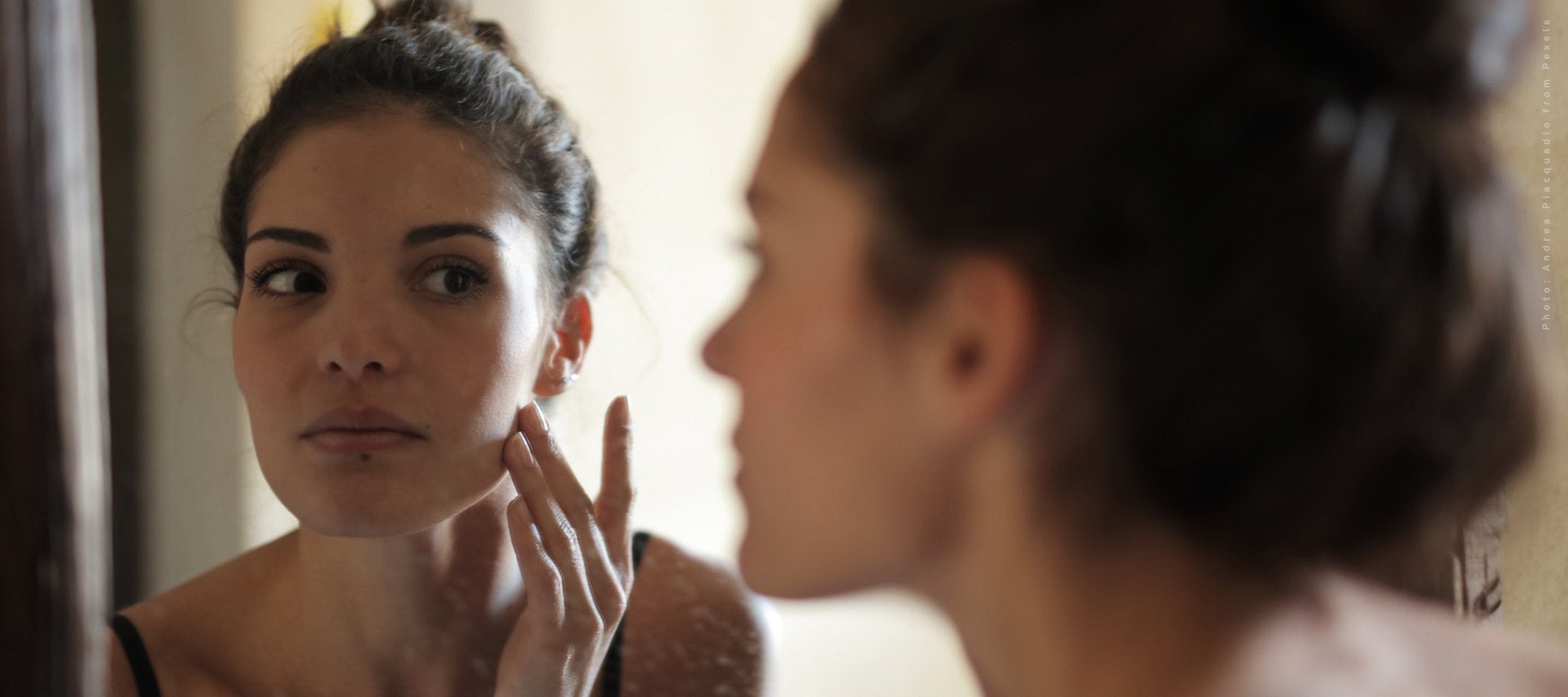 A woman with dark hair applying a skin care product to her cheek while looking intently at her reflection in a mirror.