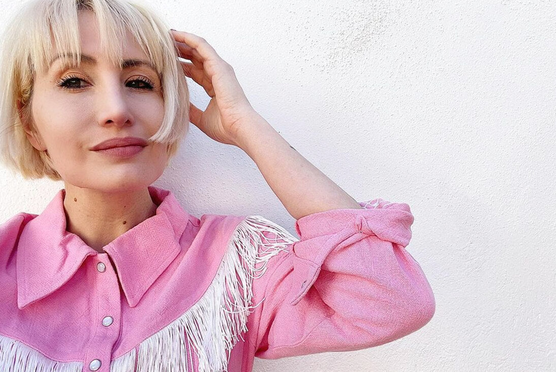 A blonde woman in a pink Western-style shirt with white fringe, posing with one hand in her hair against a plain white wall.