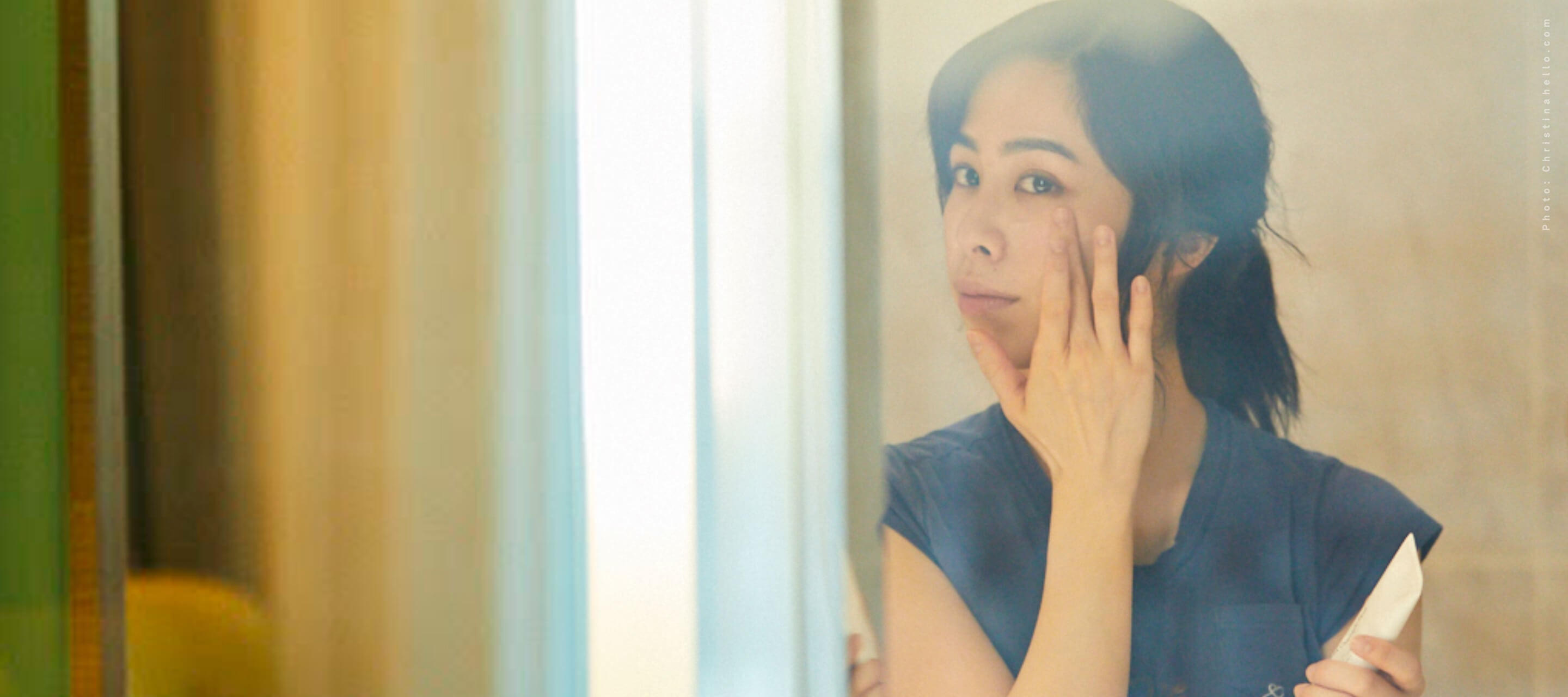 A woman with dark hair pulled back looks into a mirror while applying skincare to her cheek in a sunlit room.