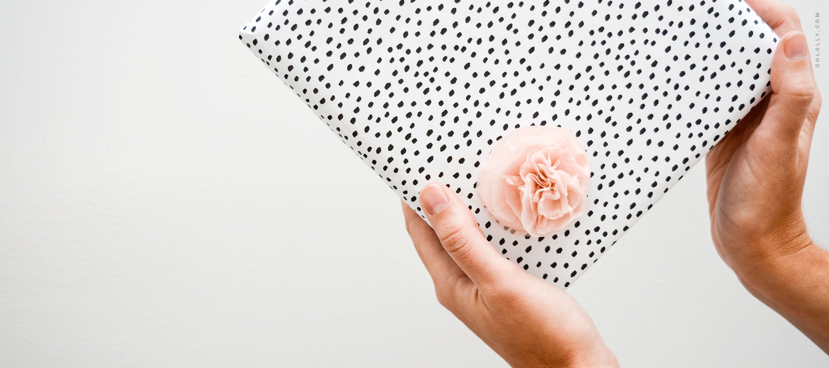 Two hands holding a gift wrapped in white paper with black dots, decorated with a small pink flower.