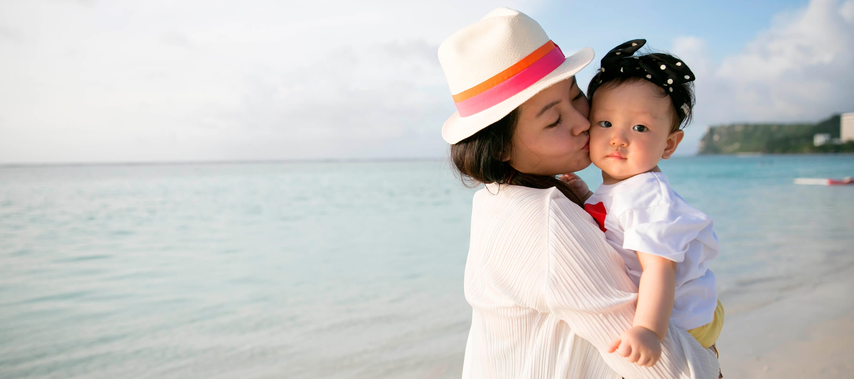 A woman wearing a sun hat kisses a young child on the cheek while standing on a beach with the ocean in the background.