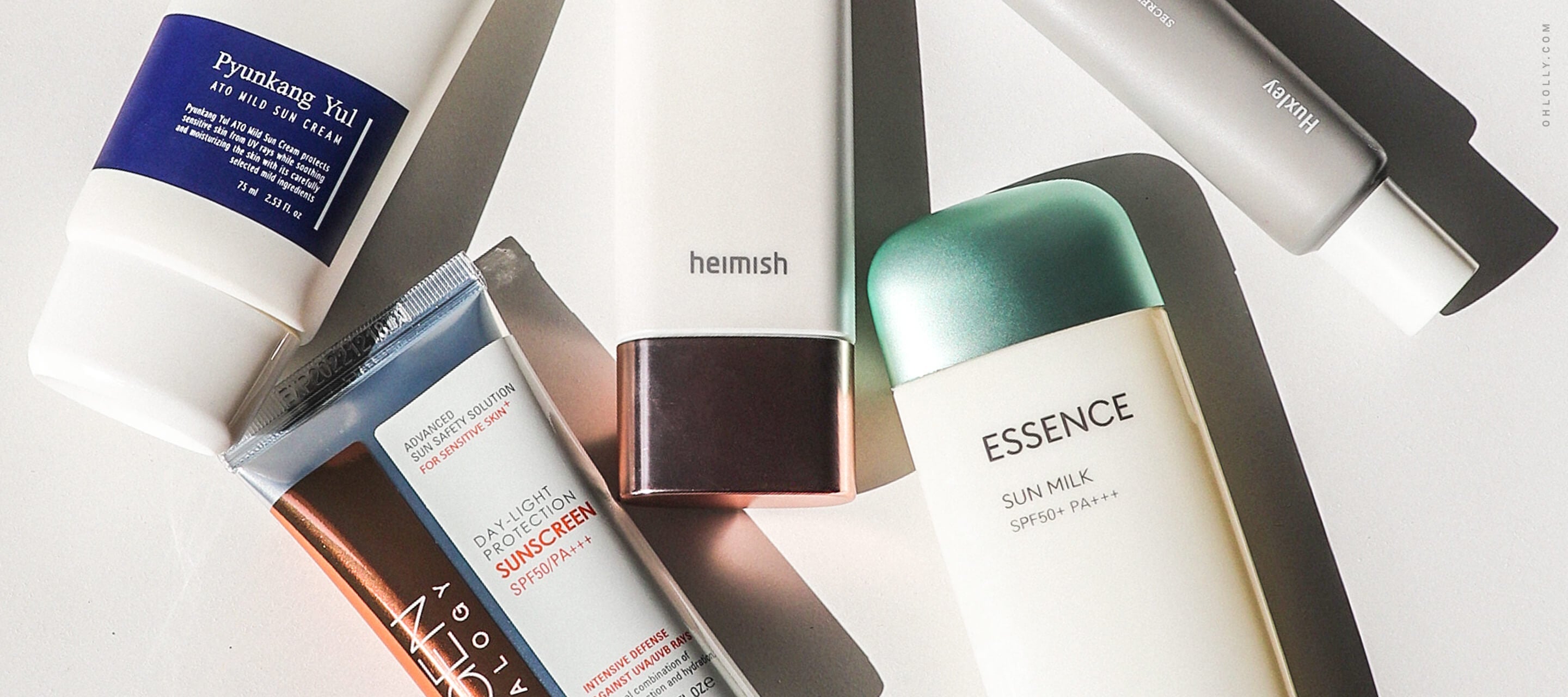 A flat lay arrangement of five different tubes of sunscreen, including brands like Pyunkang Yul, Neogen, and Missha, angled across a white surface with shadows.