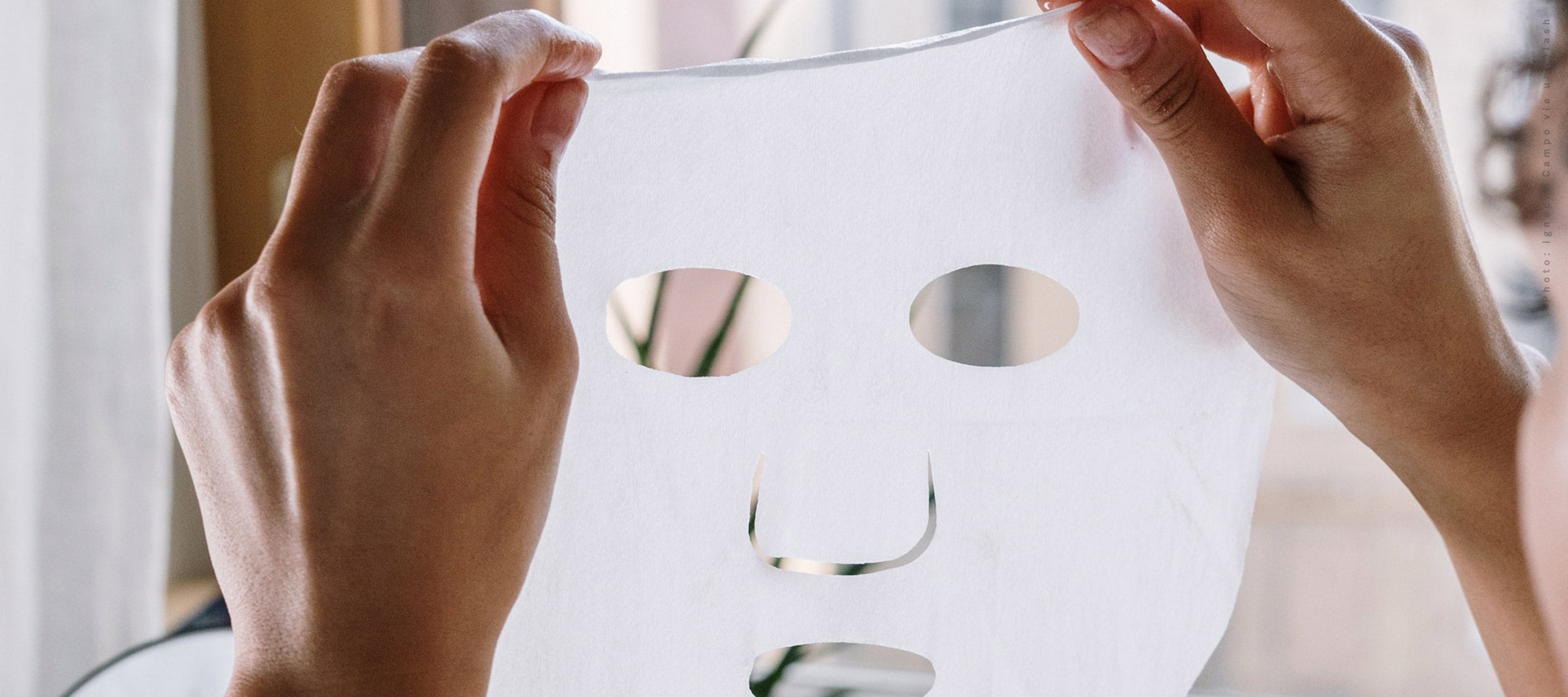 Hands holding up a white sheet mask against a brightly lit window, symbolizing relaxation, self-care, and an easy step in a K-Beauty routine.