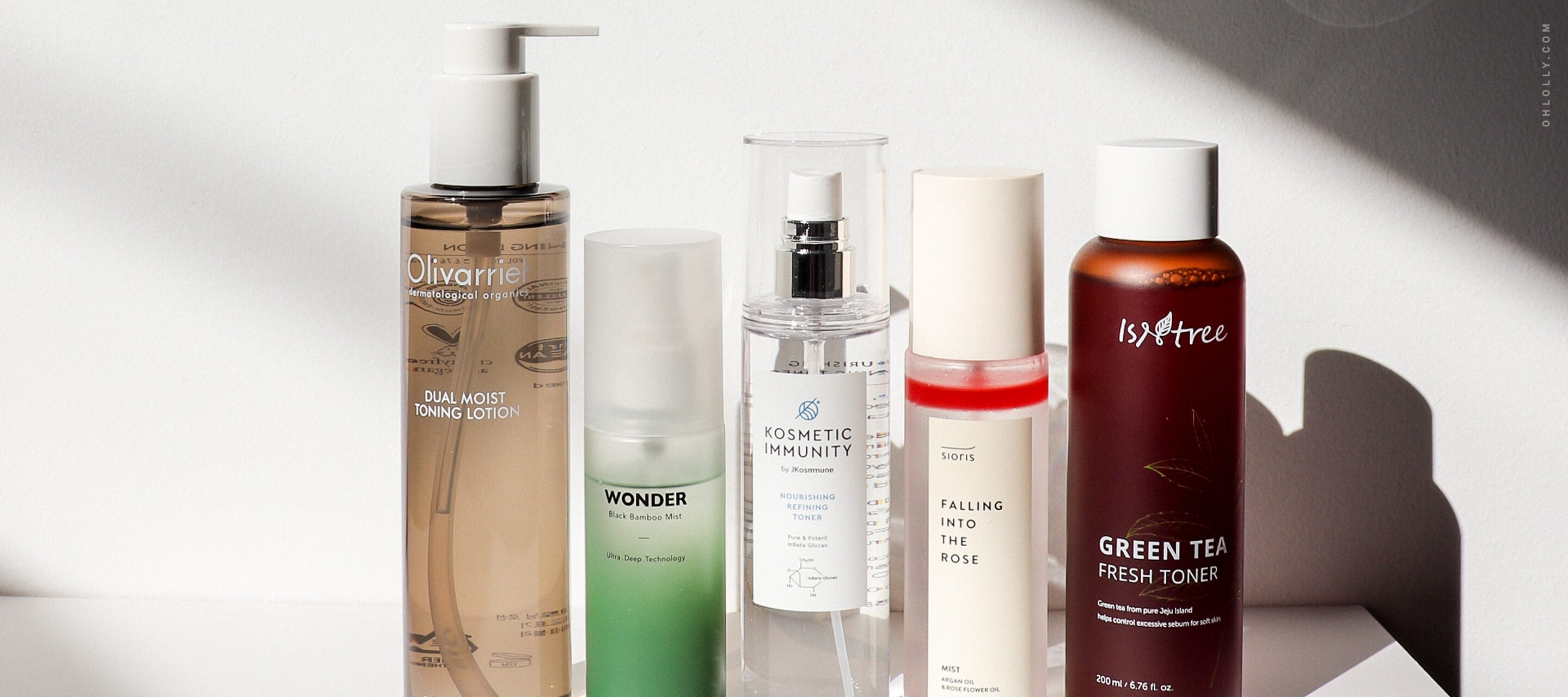 A row of five diverse skincare bottles of various sizes and colors, including a tall brown pump bottle and a green mist bottle, set against a white background with sharp shadows.