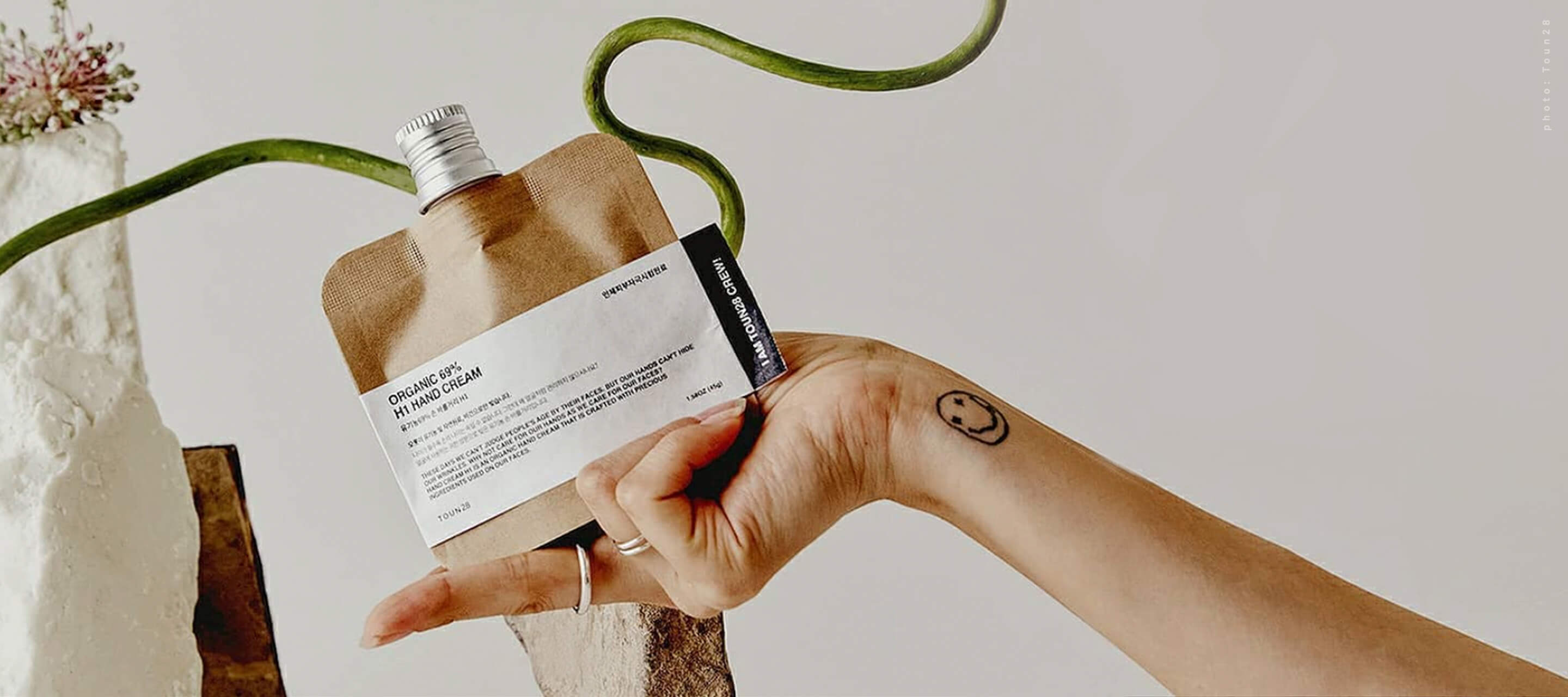 A person's hand holding a Toun28 sustainable skincare pouch against a white background with a green vine, featuring a small smiley face tattoo on the wrist.