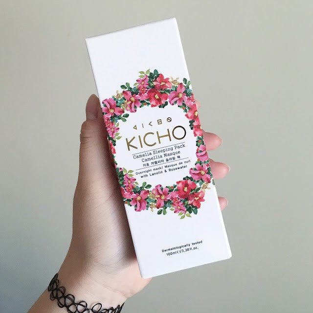 A hand holding a white box of Kicho Camellia Sleeping Pack, featuring a decorative ring of pink and red roses on the front.