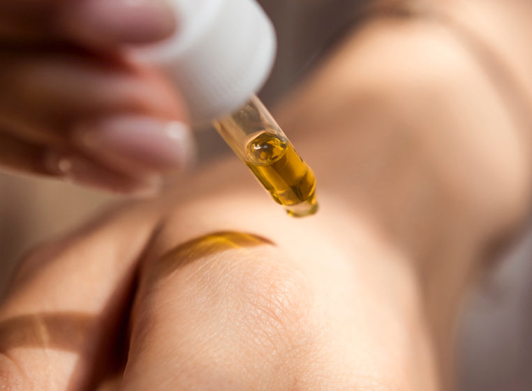 Close-up of a person's hand with a drop of golden oil being applied from a glass dropper.