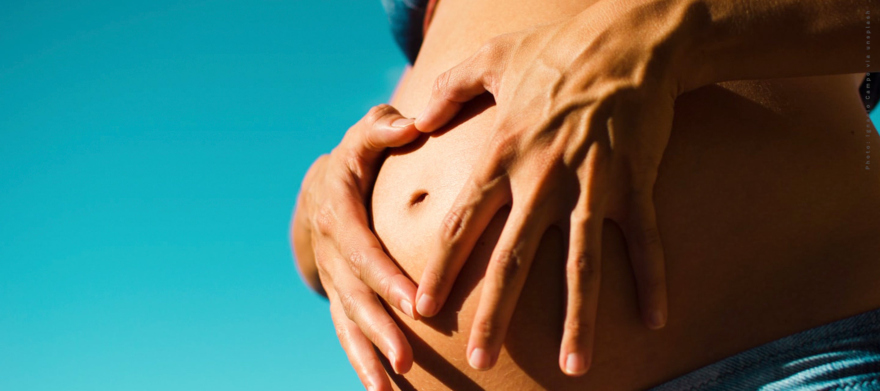 A person making a heart shape with their hands over their pregnant belly against a bright blue sky, symbolizing maternity, love, and prenatal care.