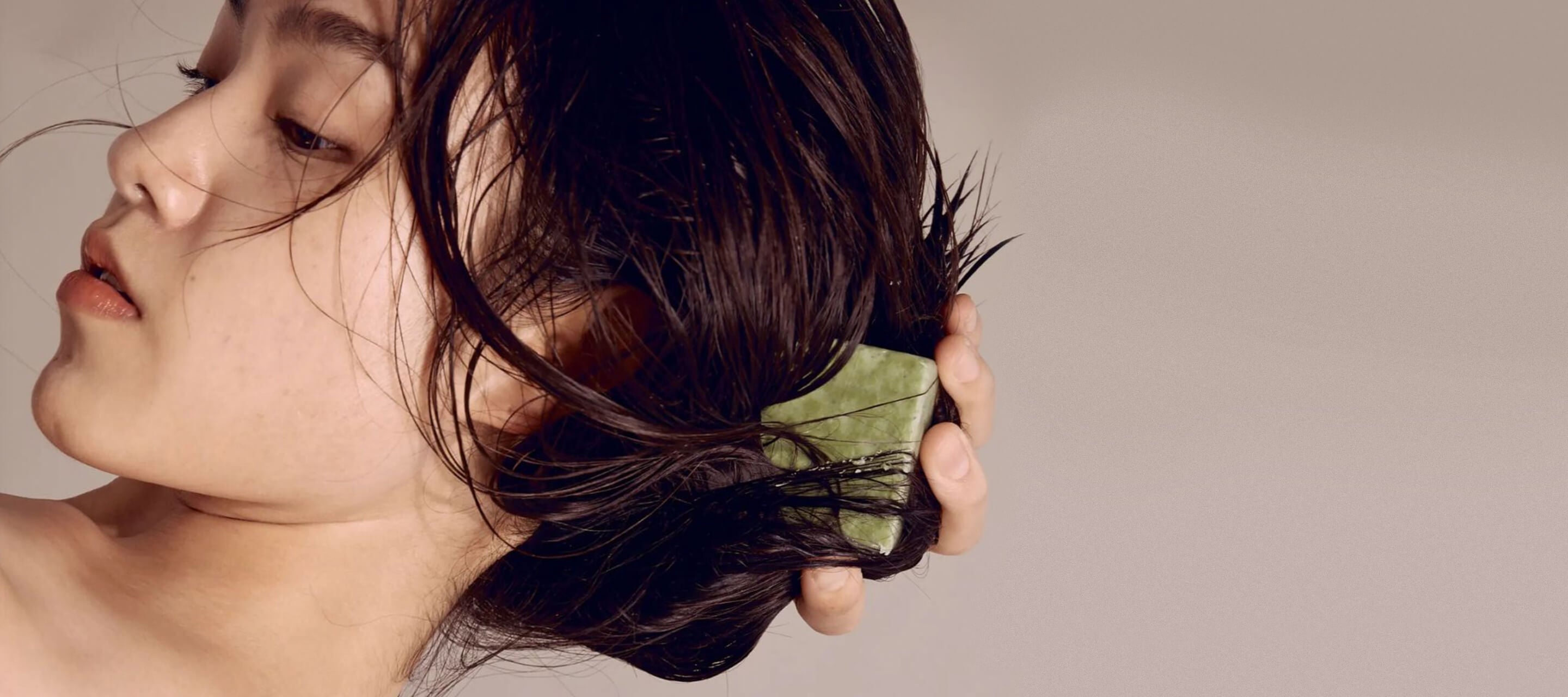 A woman with dark hair uses a green bar soap to wash her hair, shown in a side profile view against a neutral background.
