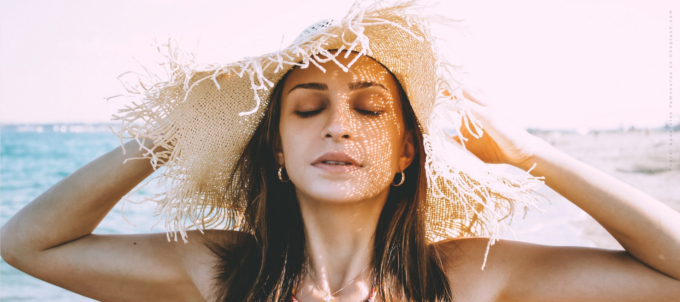 Woman in a straw hat with eyes closed on a sunny beach, highlighting sun protection and skincare.