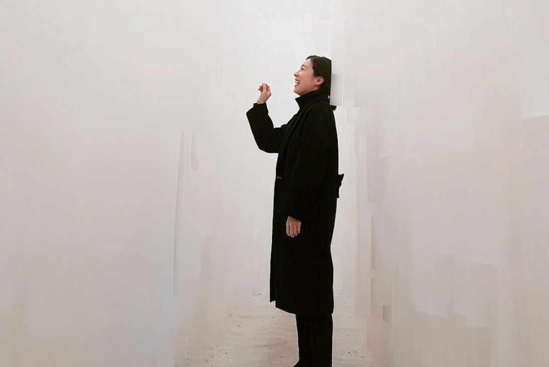 A woman wearing a long black coat stands in profile against a plain white wall, smiling and looking upward with one hand raised near her chin.