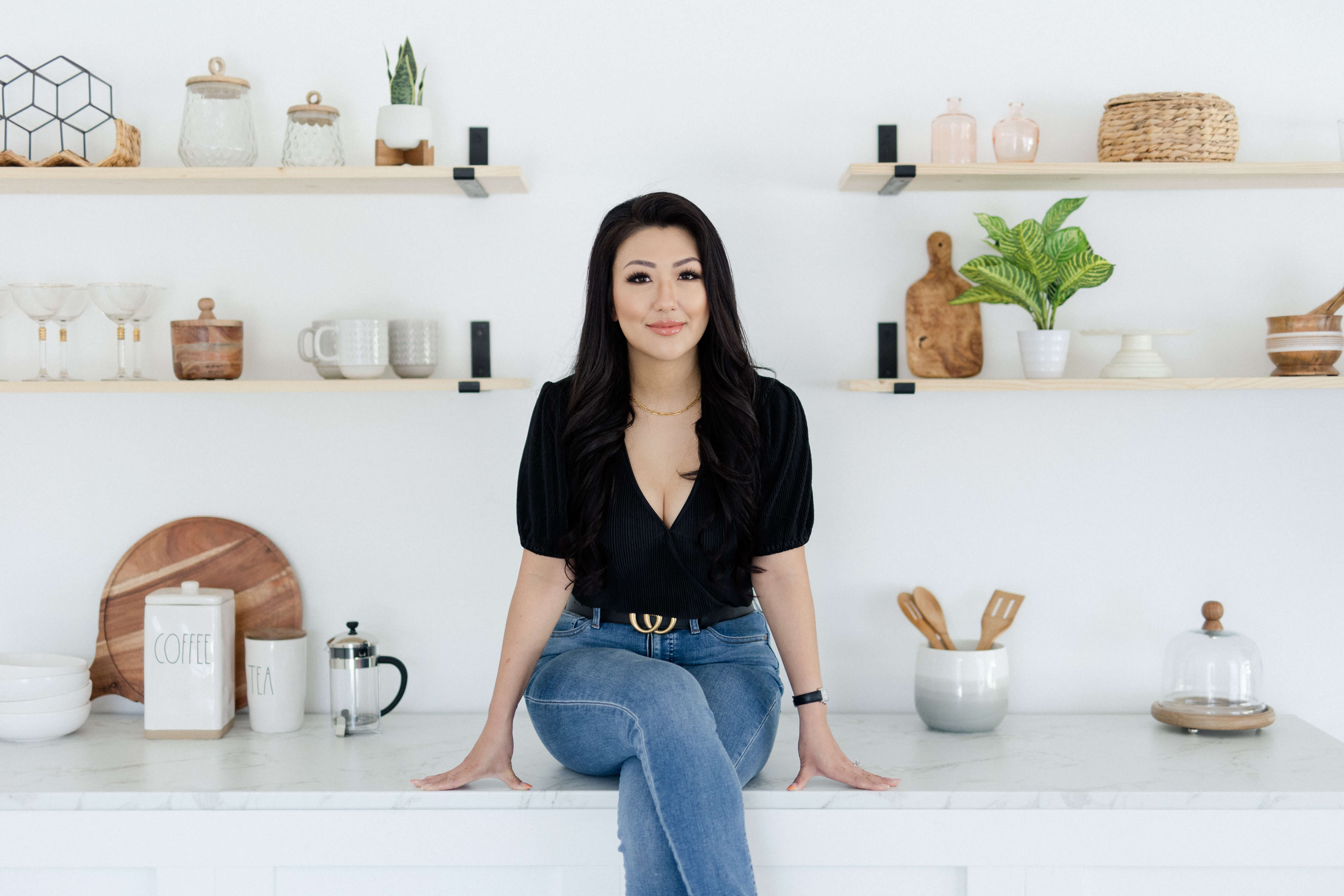 Sandra Lee, with long dark hair and a black top, sitting on a white kitchen counter in front of open shelving decorated with plants and kitchenware.