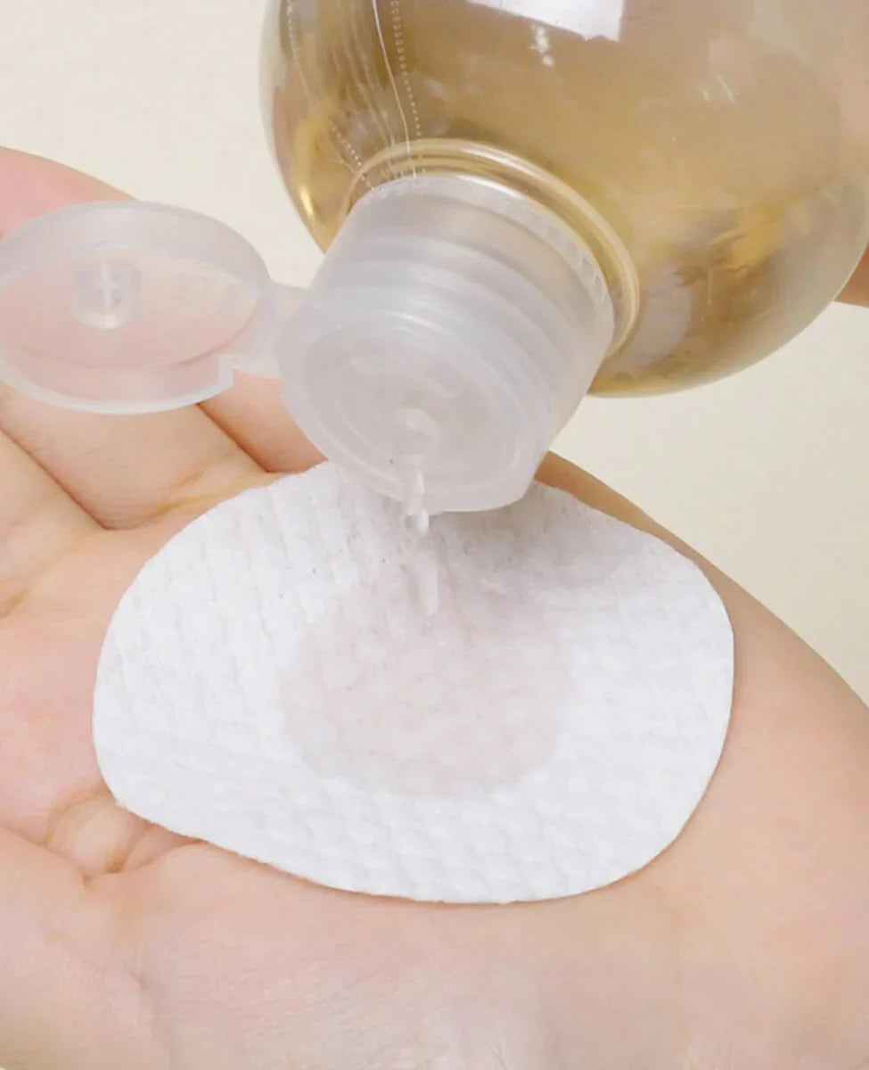 A person's hand holding a round cotton pad while a clear liquid is being poured onto it from a plastic bottle.
