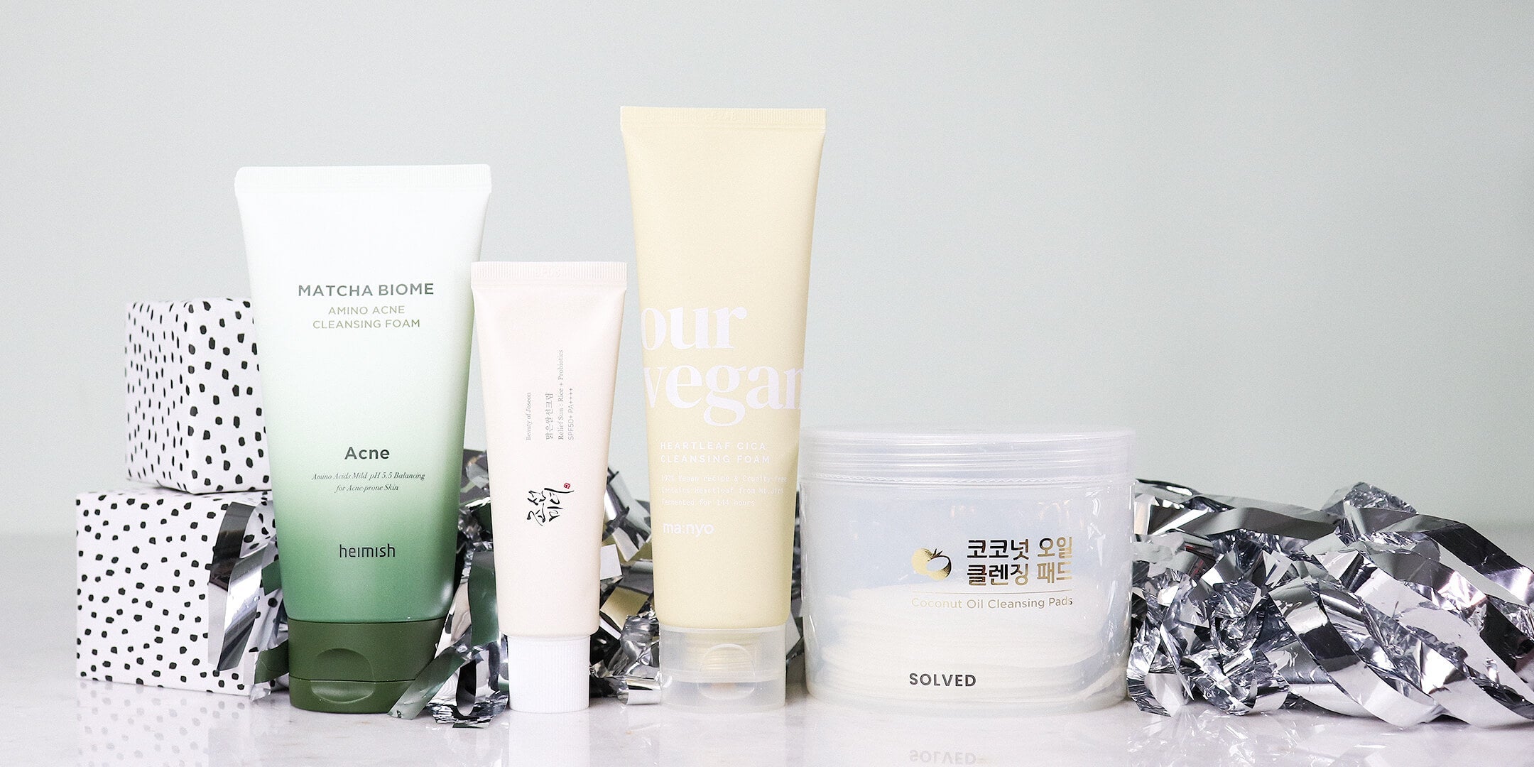 Various K-beauty cleansing products, including tubes of foam cleanser and a jar of cleansing pads, arranged with festive silver tinsel and polka-dot gift boxes.