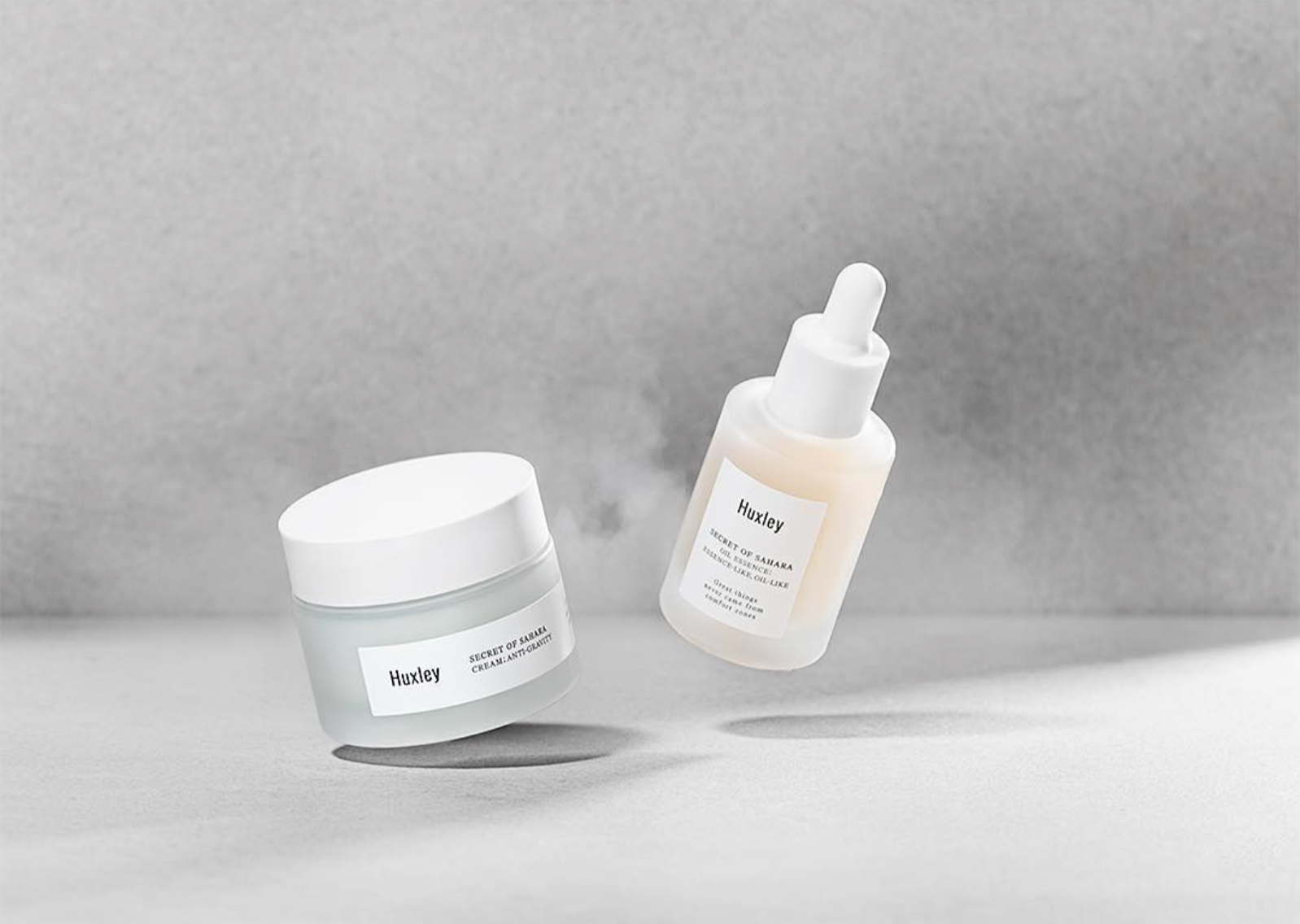 Two Huxley skincare products—a white cream jar and a frosted glass dropper bottle—floating against a solid grey background.