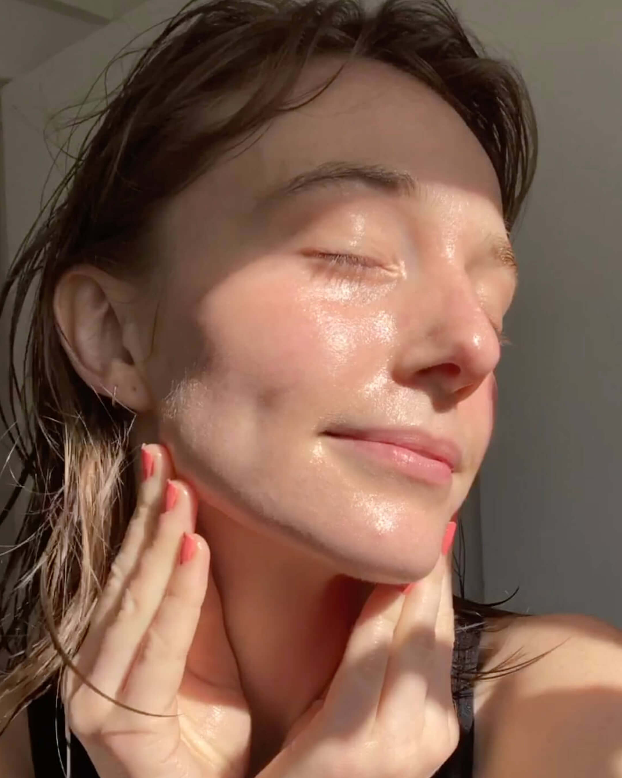 A close-up of a person's face with glowing skin, with their hands gently touching their jawline in natural sunlight.