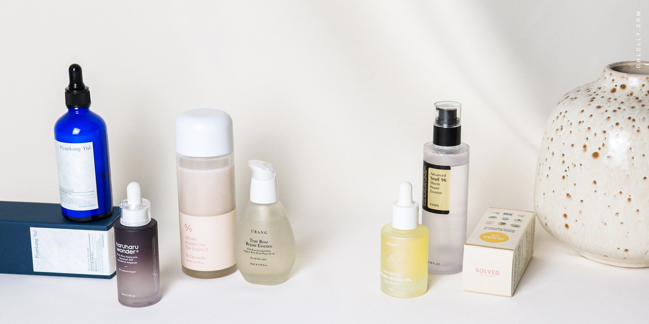 A collection of six K-beauty skincare products, including various serums and toners in bottles, arranged on a white surface next to a speckled ceramic vase.