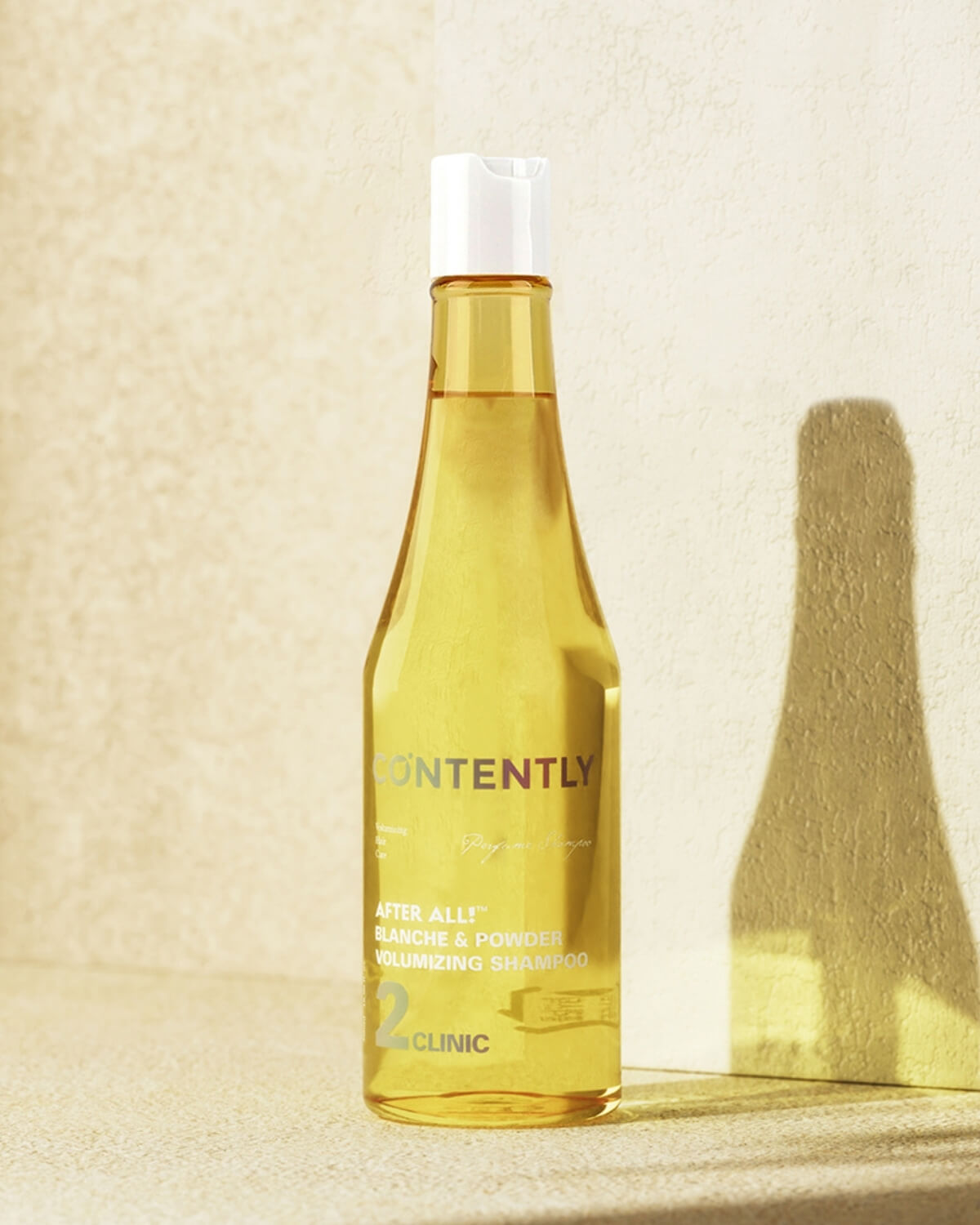 A transparent yellow bottle of CONTENTLY CALIFORNIA "After All! Blanche & Powder Volumizing Shampoo" with a white cap is standing on a light beige surface. A strong, dark shadow of the bottle is cast to the right on the textured wall in the background.