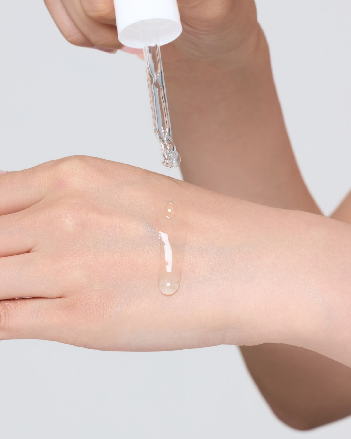 A close-up of a hand using the dropper to dispense a thick, clear drop of the serum onto the back of the other hand, demonstrating the product's texture.