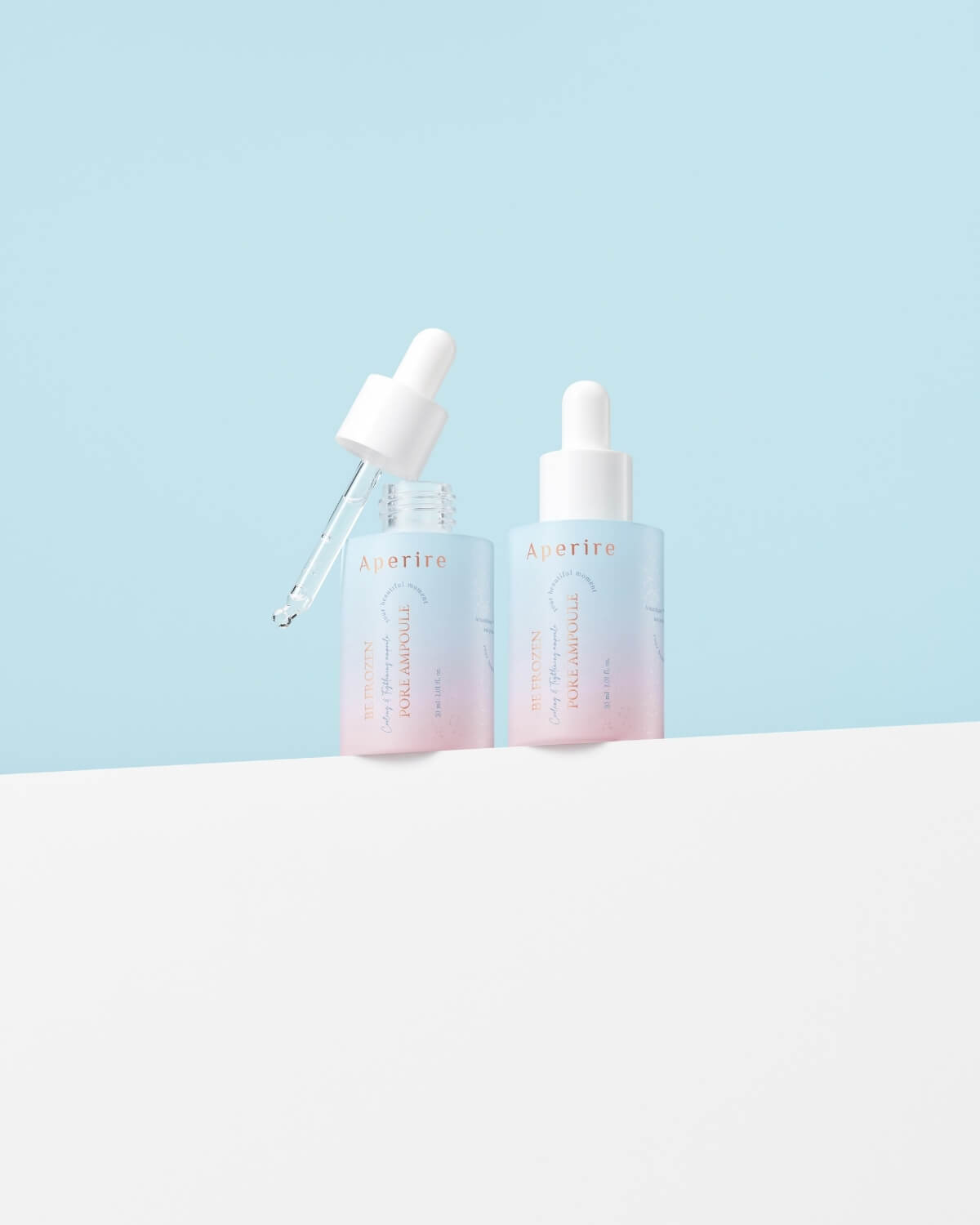 Two ombre ampoule bottles sit side-by-side on a white surface with a light blue background. One bottle has its dropper removed, suspended just above the opening.