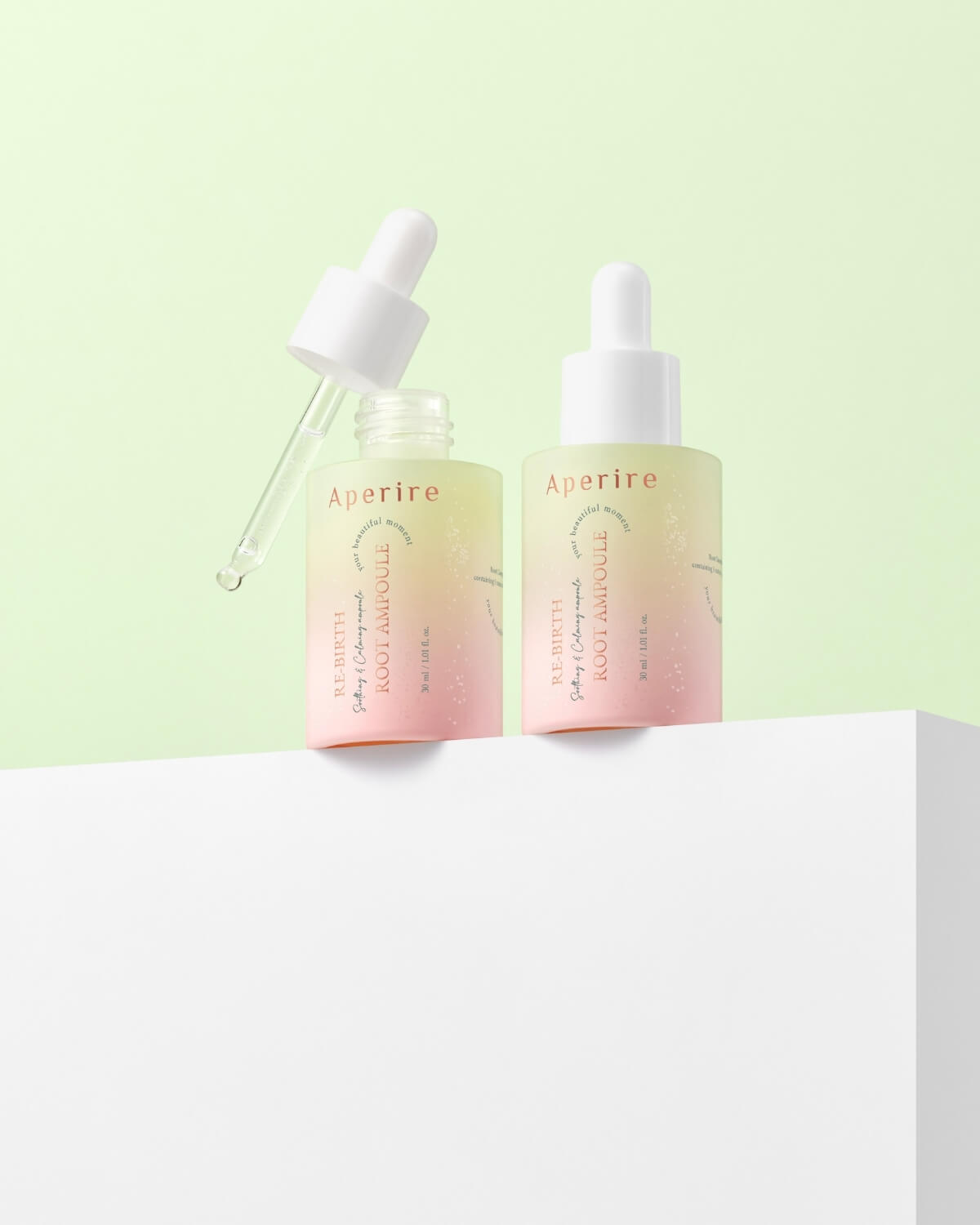 Two bottles of Aperire Re-birth Root Ampoule are displayed on a white platform against a soft green background. The bottle on the left has its white dropper removed and dripping a drop of the serum, while the bottle on the right is closed.