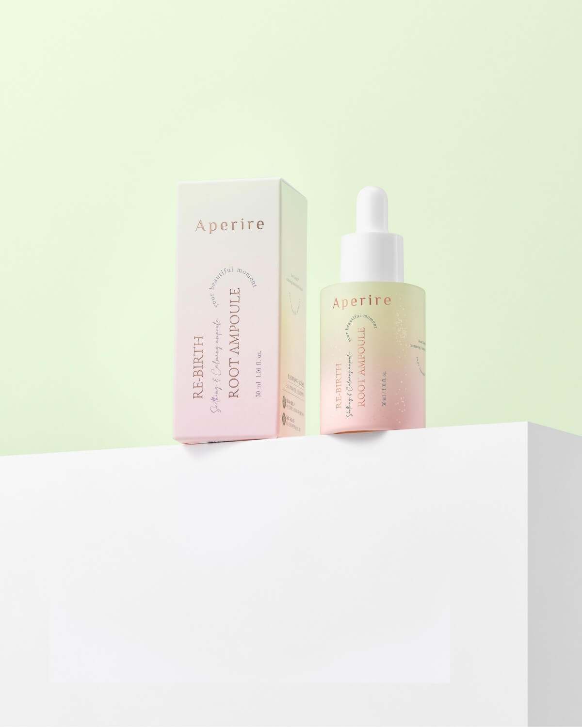 A pink and yellow ombre bottle of Aperire Re-birth Root Ampoule in its matching pale pink box, resting on a white geometric platform against a soft green background. The bottle's label reads "Re-Birth Root Ampoule Soothing & Calming Ampoule."