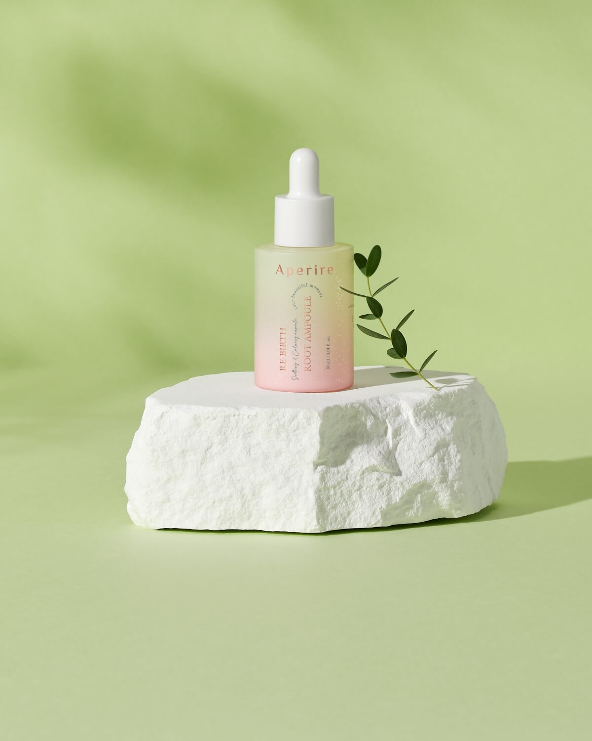 The Aperire Re-birth Root Ampoule bottle sits on a rough, white stone platform. A small green sprig of leaves is placed next to the bottle. The background is a soft green with subtle shadows creating a natural, serene aesthetic.