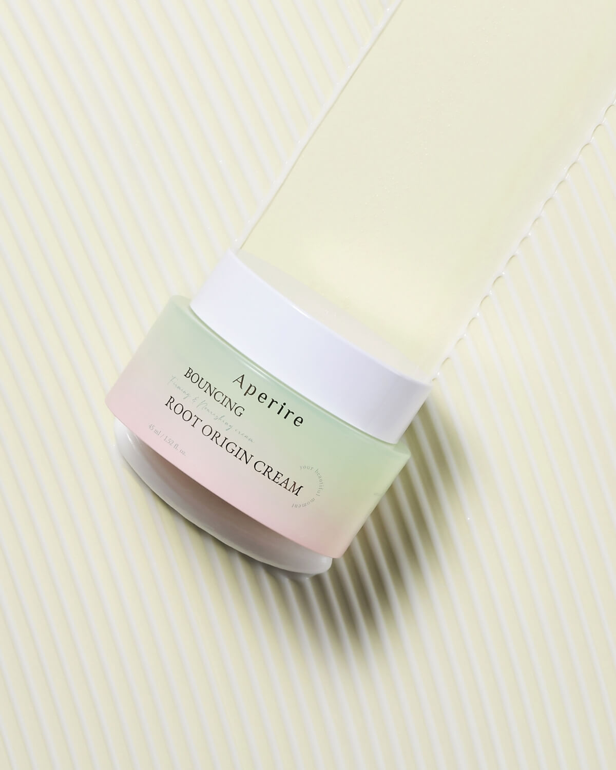 A top-down, artistic shot of the Aperire Bouncing Root Origin Cream jar resting on a ribbed, pale yellow surface. The jar is tilted, and a streak of the cream texture is visible extending above the jar.