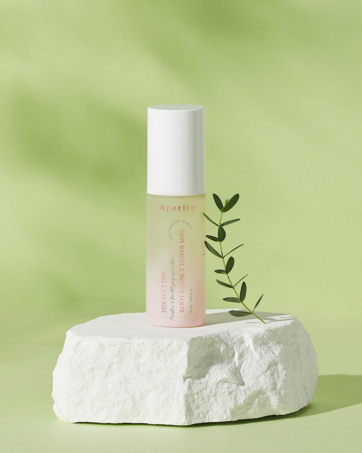 A bottle of Aperire Miracle Day Root Essence Toner Mist resting on a textured white stone block, with a small green sprig lying beside it. The background is light green.