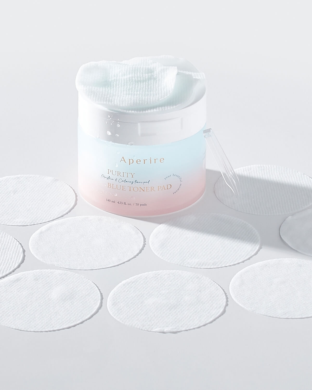 The open container of blue toner pads is displayed with several individual round white pads laid out around it on a white surface. A clear plastic tweezer is resting nearby.