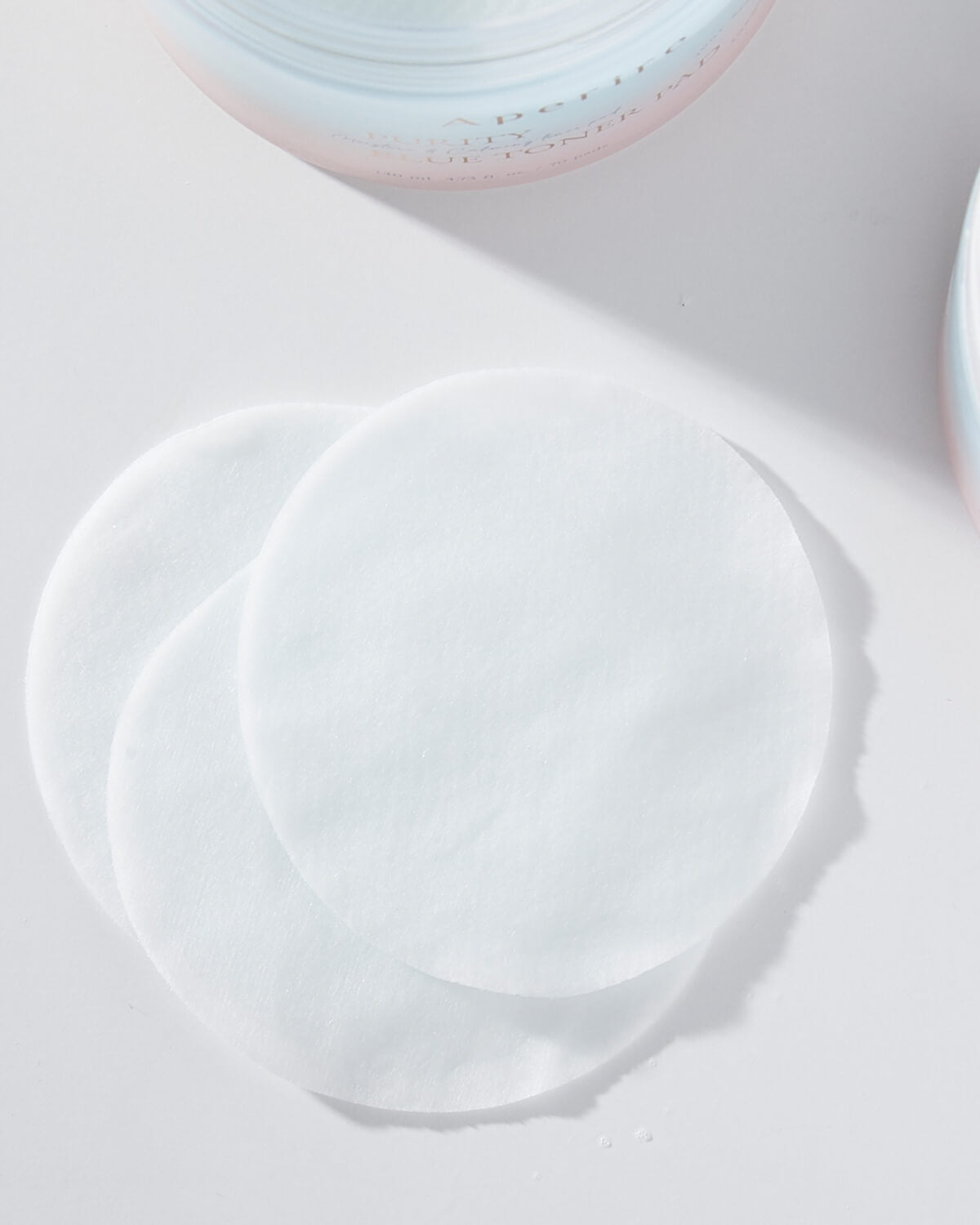 A close-up, high-angle shot of three slightly overlapping white, round toner pads lying on a bright white surface, with a hint of the container visible at the top.