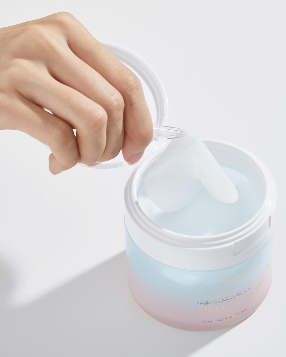 A hand lifts the protective inner seal from the open container of blue toner pads, revealing the pads submerged in a blue-tinted liquid inside the ombre jar.