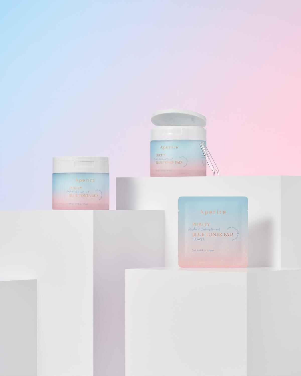 A compositional shot showing the large, open toner pad jar and a closed jar displayed on separate white steps, alongside an individual travel sachet of the Aperire Purity Blue Toner Pad, all against a colorful blue and pink background.