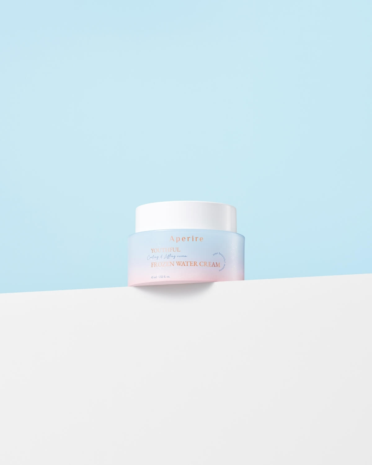 The cream jar with its blue-to-pink ombre design sits squarely on the edge of a clean white surface against a solid light blue background.