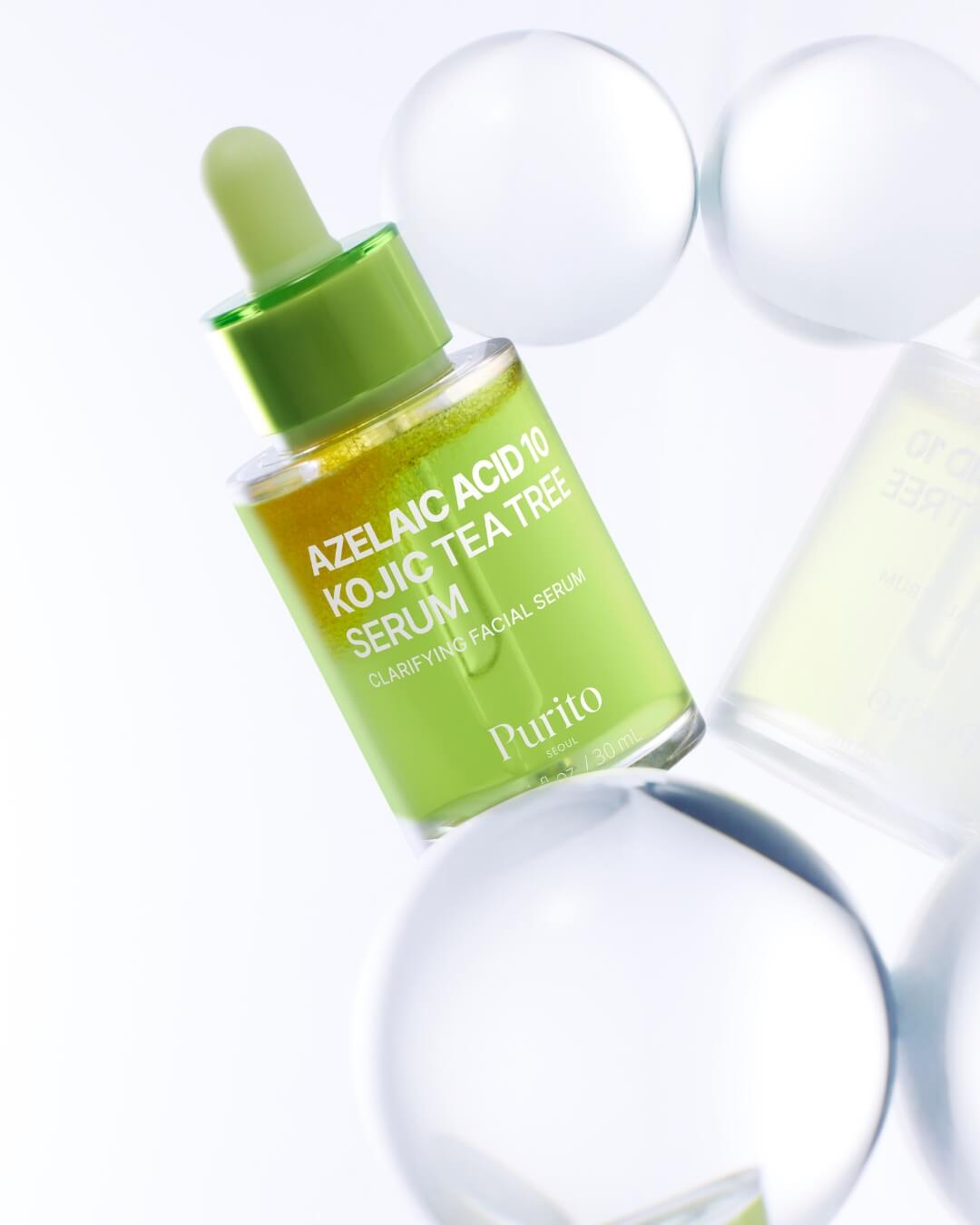 A dynamic studio shot of the Purito Azelaic Acid 10 Kojic Tea Tree Serum bottle tilted and surrounded by several blurry white glass spheres on a white background. The bottle's vibrant green and yellow bi-phase liquid is the focal point.