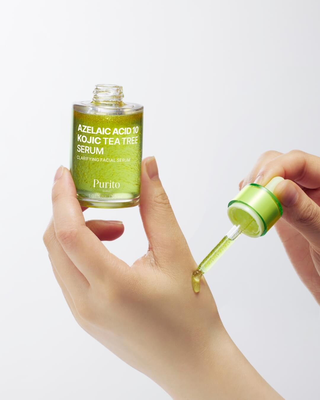 A person's hands holding the Purito Azelaic Acid 10 Kojic Tea Tree Serum. One hand holds the bottle, and the other uses the dropper to dispense a large drop of the green serum onto the back of the hand.