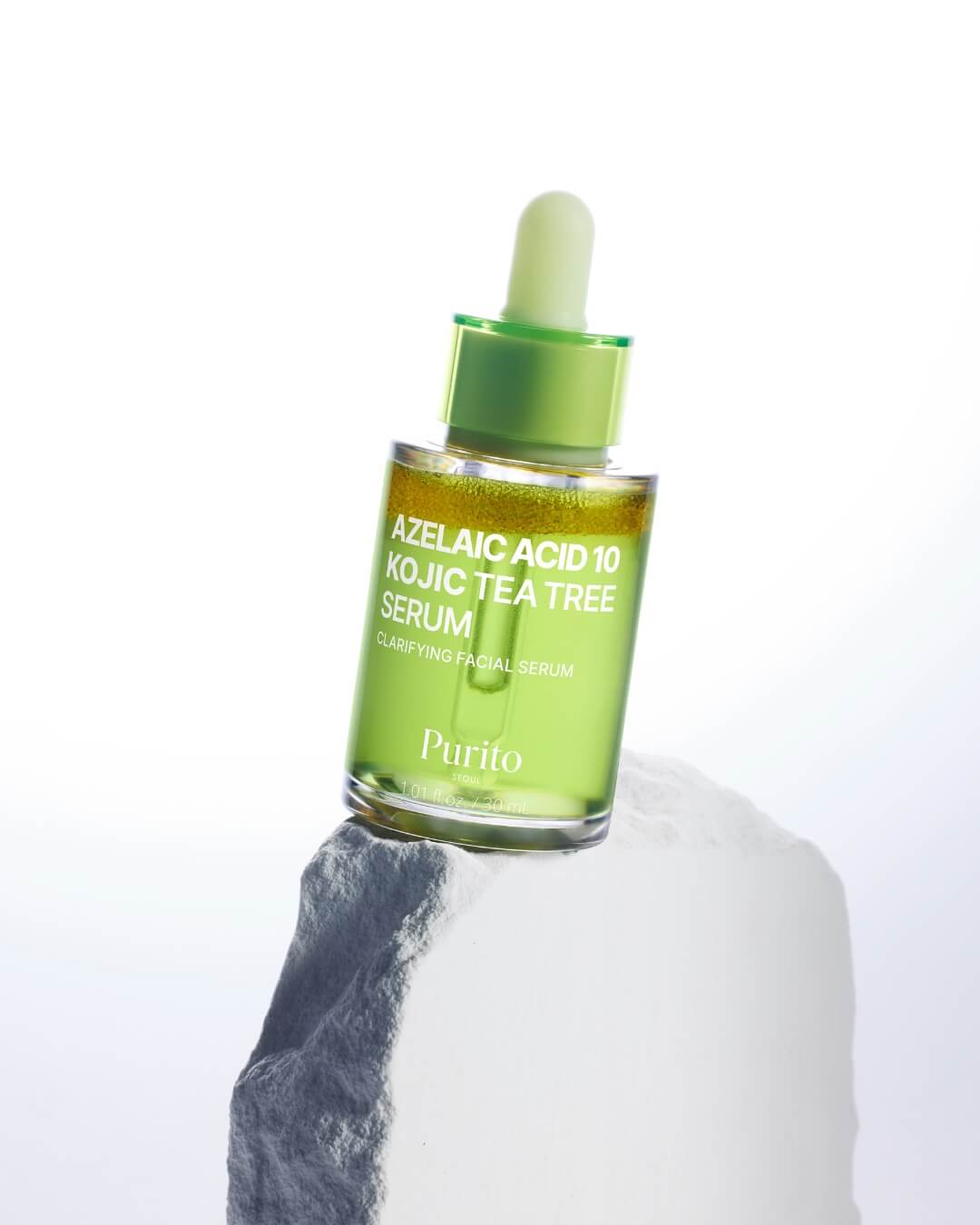 A bottle of Purito Azelaic Acid 10 Kojic Tea Tree Serum perched on a textured white rock against a bright, subtly graded background. The bottle is slightly tilted, highlighting the separate golden-yellow and lime-green layers of the serum.