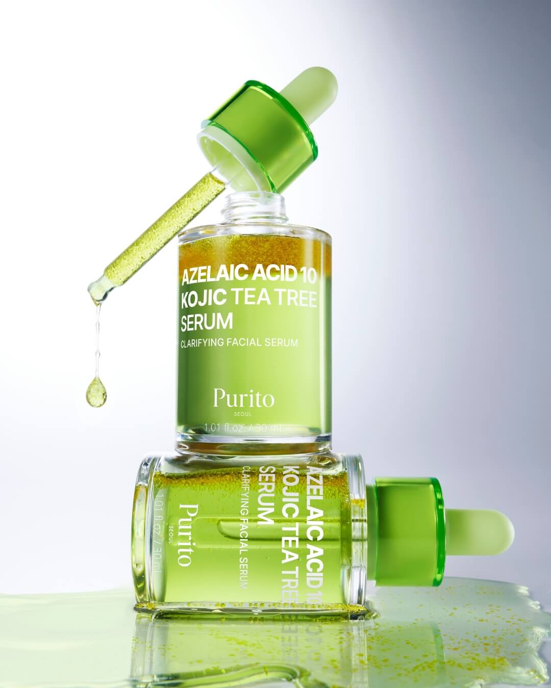 Two bottles of Purito Azelaic Acid 10 Kojic Tea Tree Serum stacked vertically. The top bottle has its dropper partially pulled out, dripping a drop of the serum, while the bottom bottle is lying horizontally on a reflective surface with spilled product.