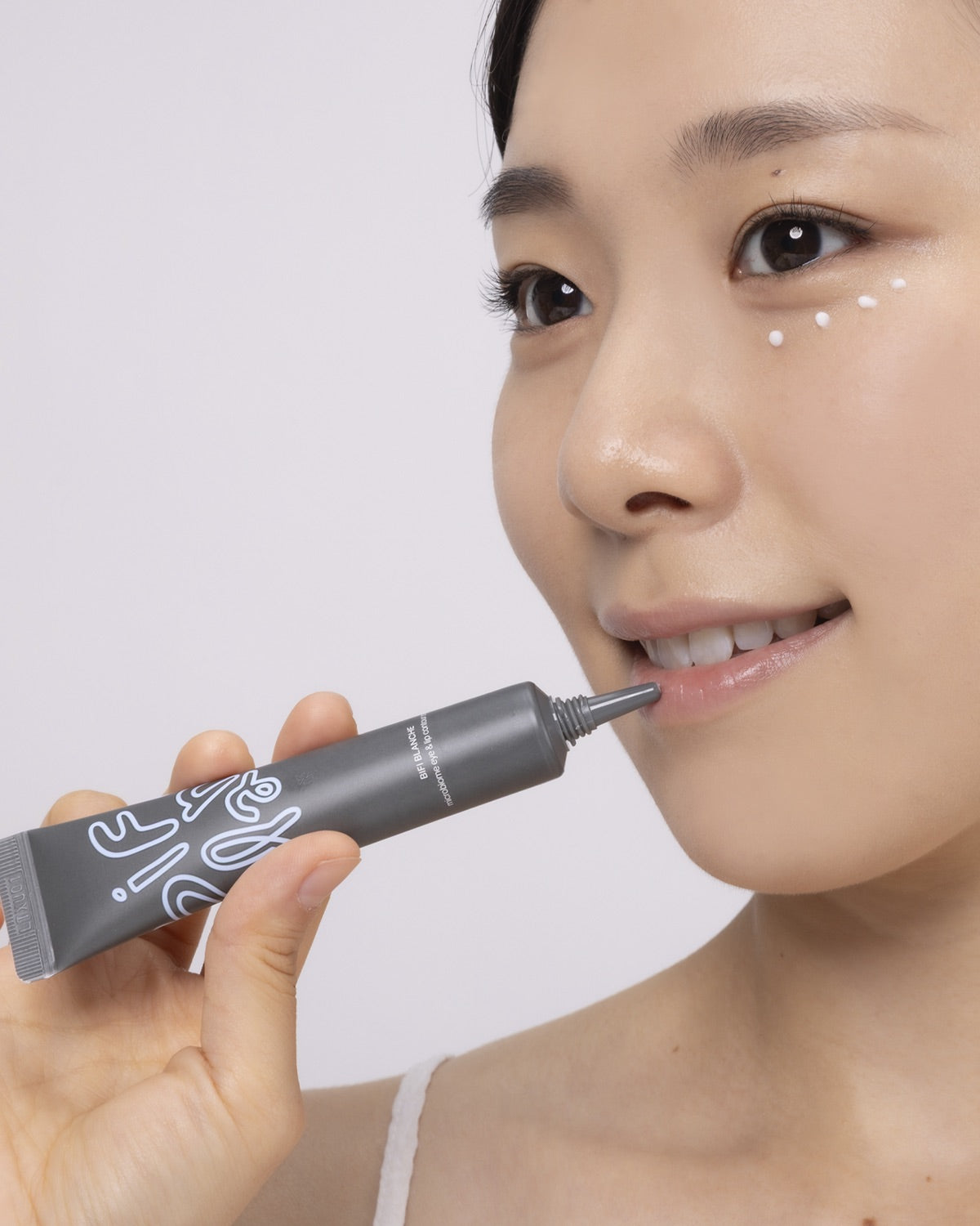 A woman applying Bifi Blanche Microbiome Eye and Lip Cream to her lips, with small cream dots under her eye.